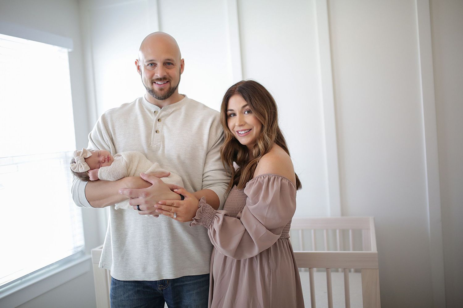 A couple in casual clothing stands by a window while holding their newborn baby.