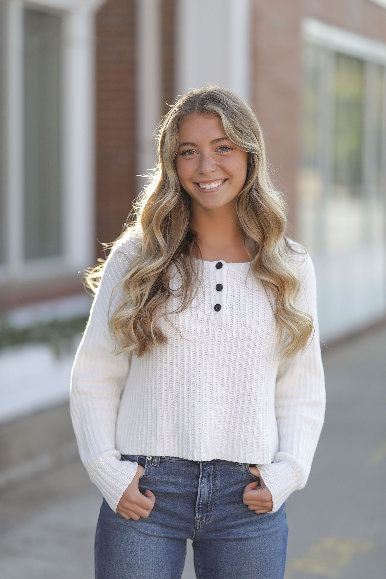 A person wearing a white ribbed henley sweater and blue jeans poses outdoors with a bright smile.