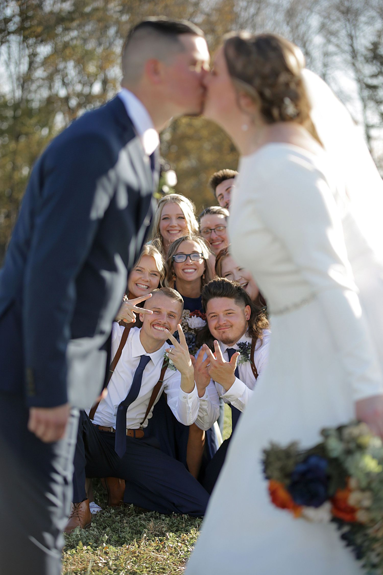 Wedding guests share excited reactions during outdoor ceremony with autumn trees in background.