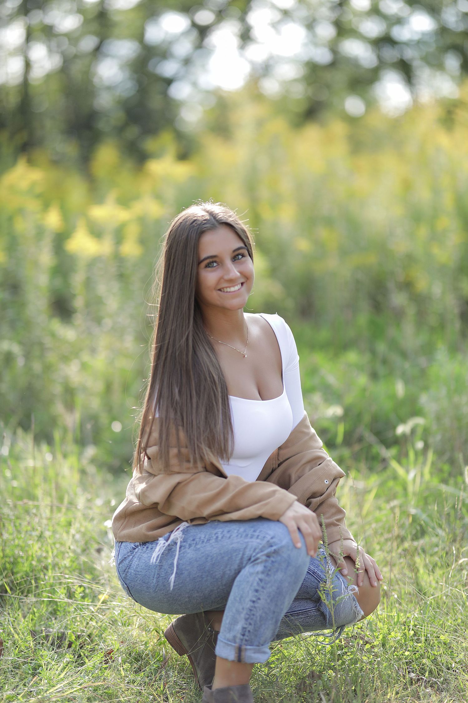 Person in white tank top and jeans crouching in a sunny meadow with yellow wildflowers and trees in the background.