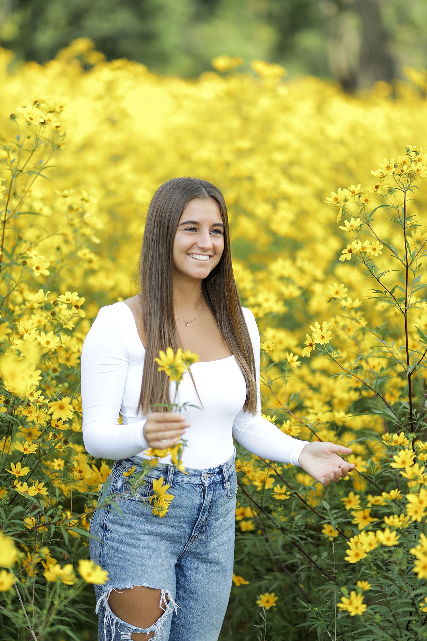 Young woman in white top and denim poses joyfully among vibrant yellow wildflowers on a sunny day.