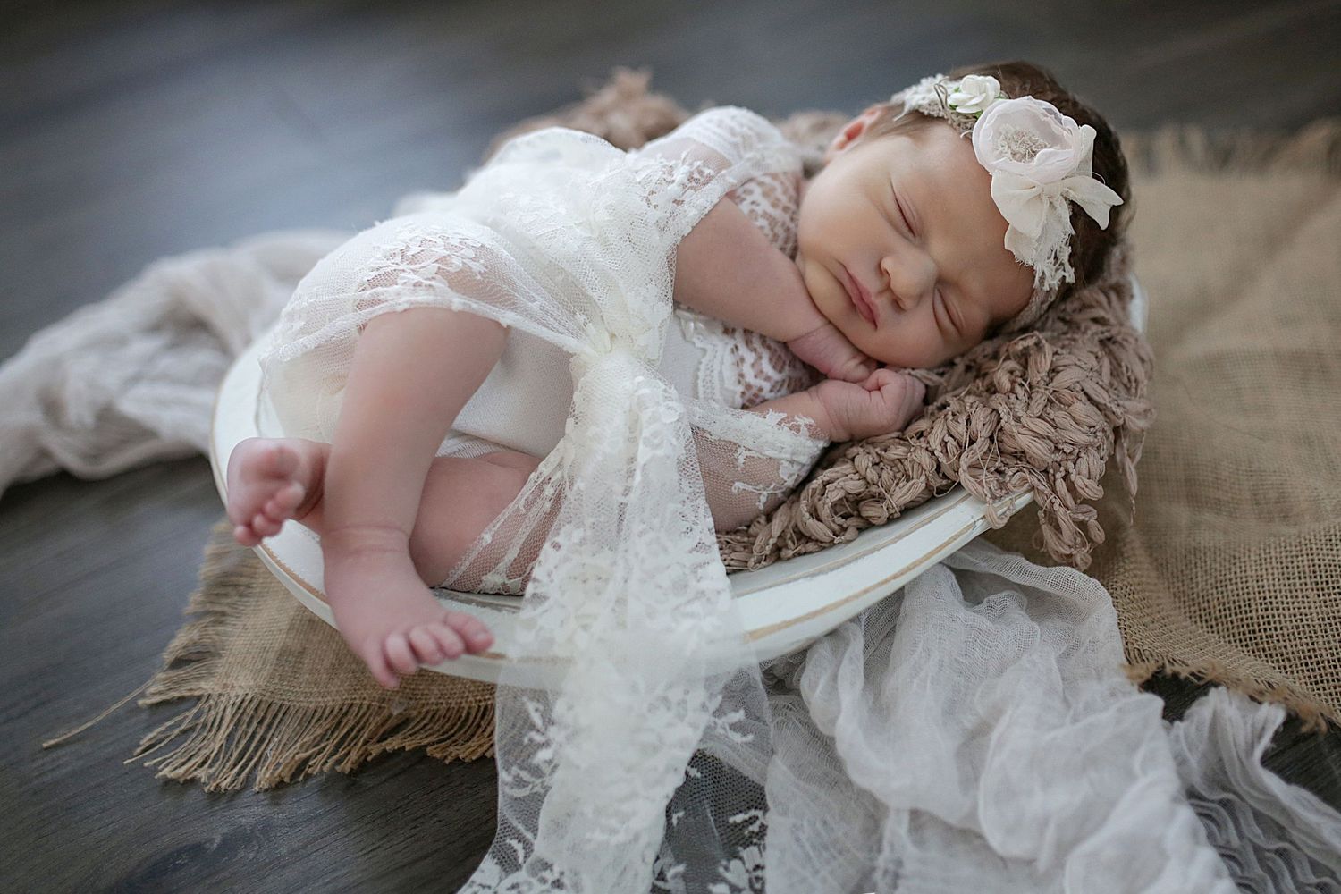 Sleeping newborn wearing delicate white lace dress and floral headband rests peacefully on textured blanket.
