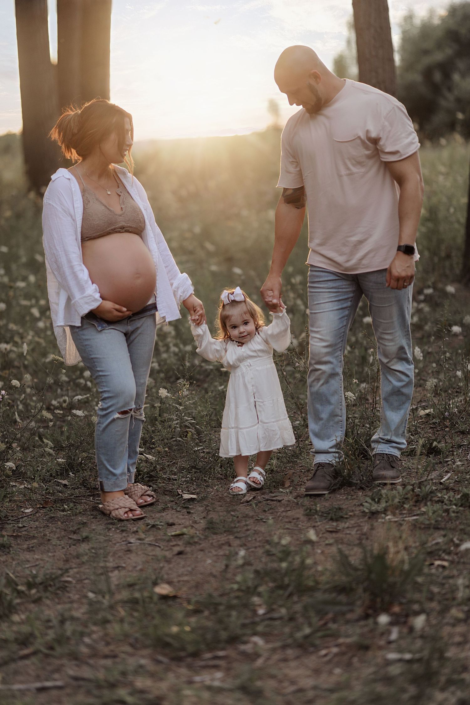 A family enjoys a sunset moment together outdoors, with a small child in a white dress walking between adults.