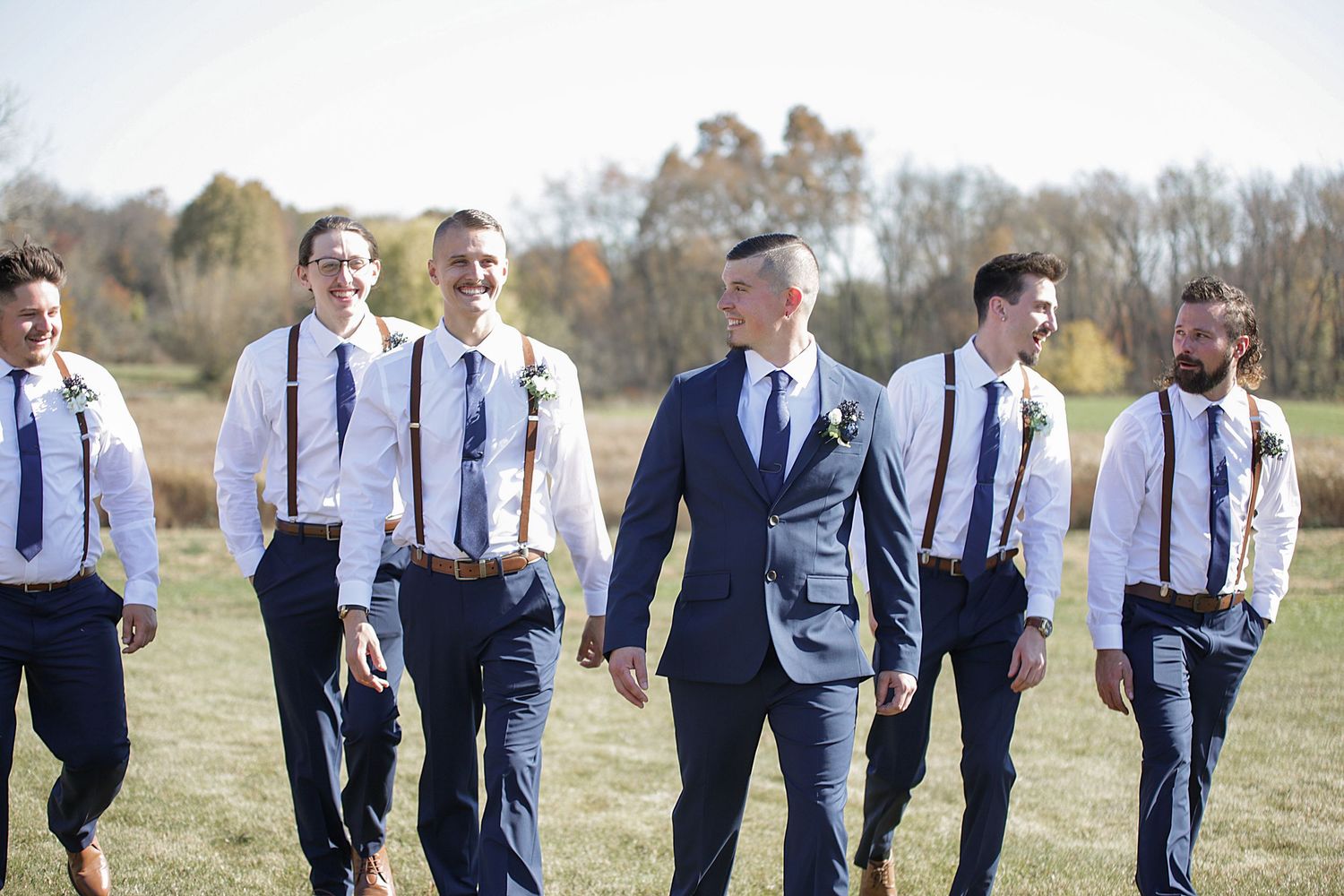 Wedding party in navy suits and suspenders walking through autumn field.