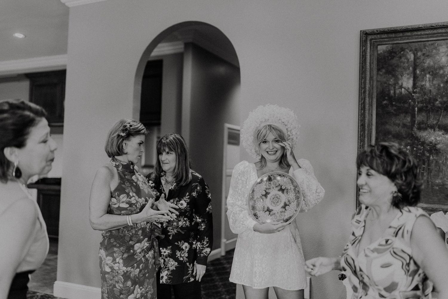 A black and white candid photo shows a group of women in fancy dresses and floral prints chatting at a social gathering.