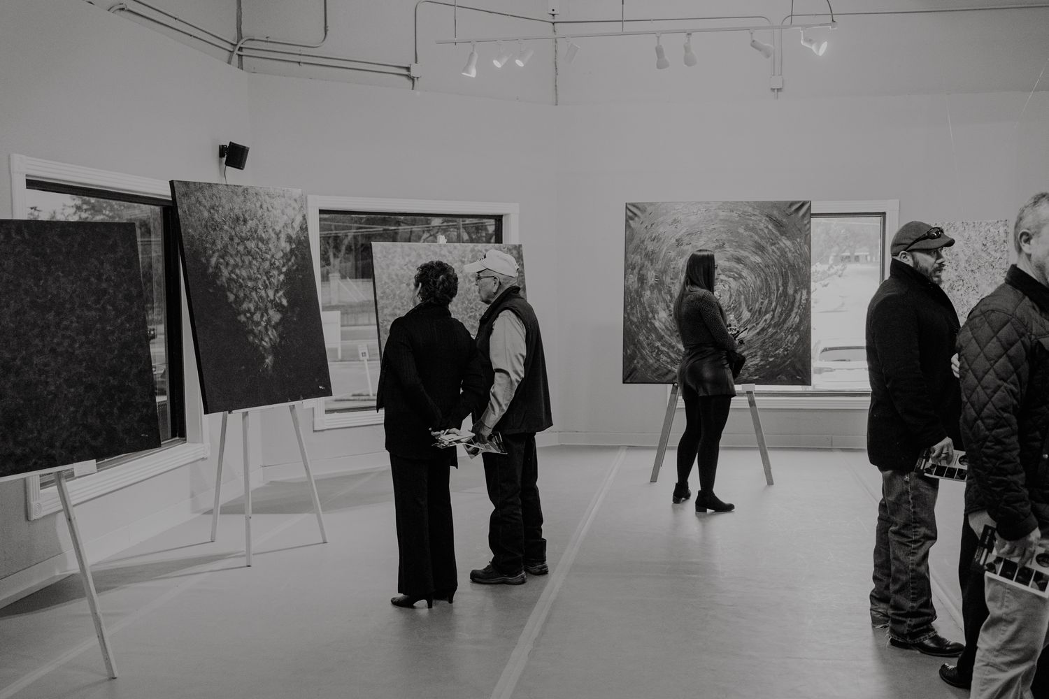 People view displayed works of art at a gallery exhibition with easels and artwork mounted on white walls.