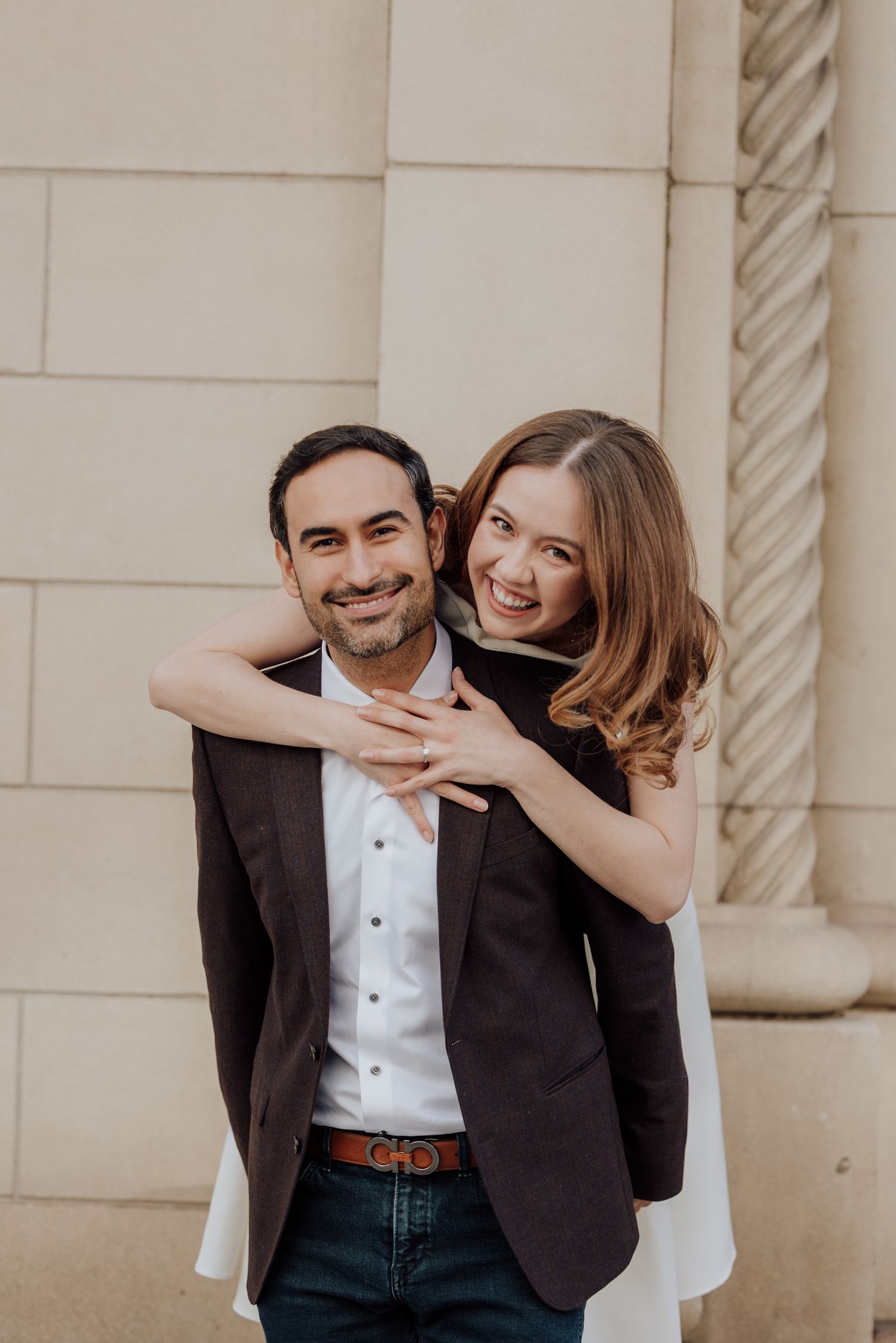 Happy couple embraces and laughs together during engagement photo session against stone wall.