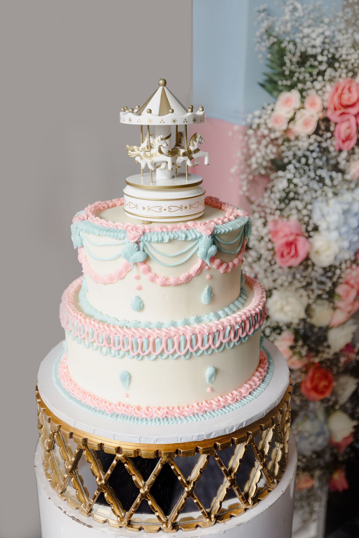 Elegant three-tiered carousel cake with pink and blue detailing topped with a white miniature merry-go-round decoration.