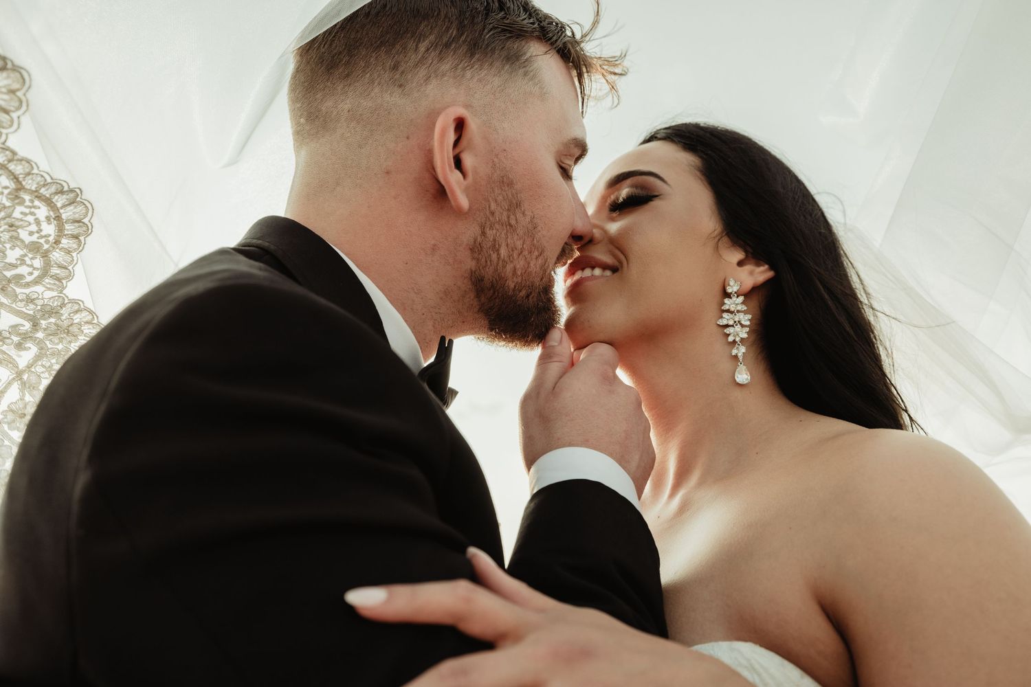 A loving couple shares an intimate romantic moment on their wedding day near sheer white curtains.