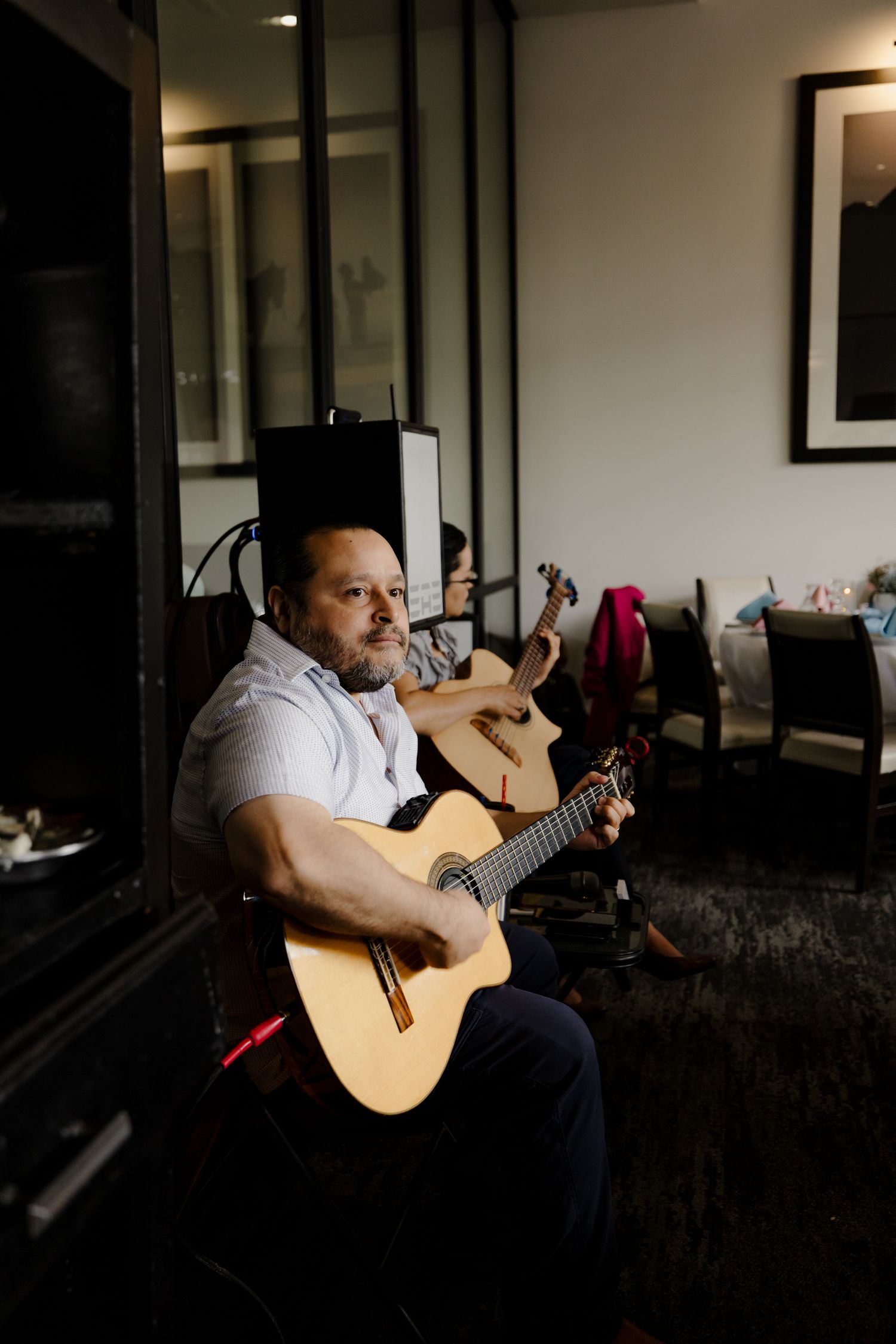A musician plays an acoustic guitar while sitting in a dimly lit room with framed artwork visible on the wall.