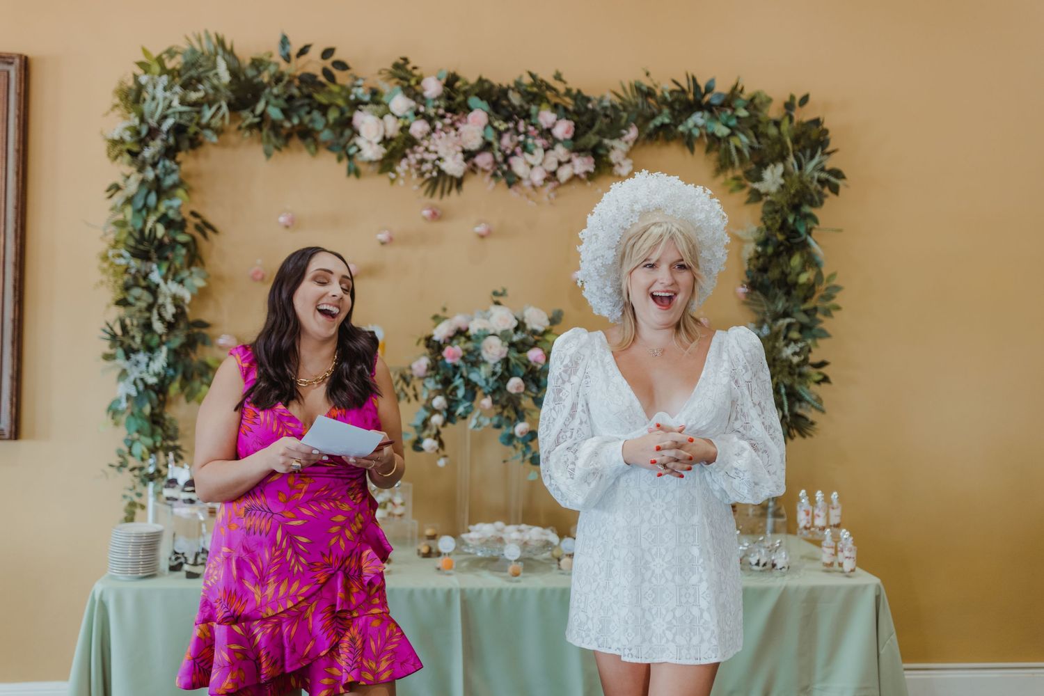 Wedding party group in elegant pink and white dresses poses together against a beige wall with floral decorations.