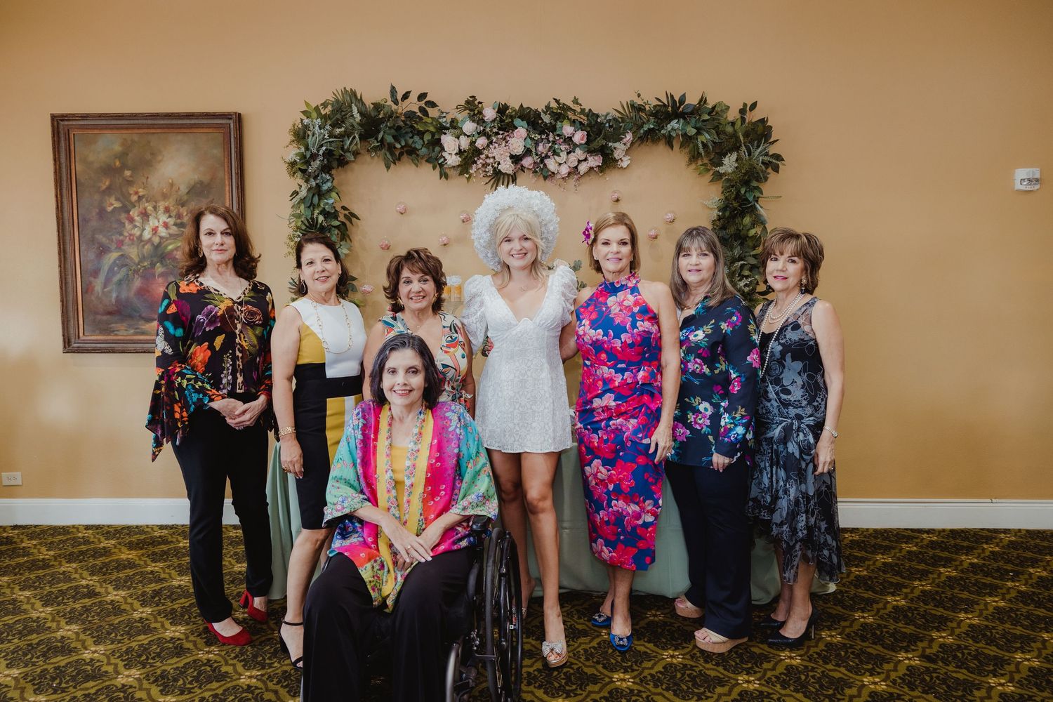 A group of people stand together and smile in front of a floral arch backdrop against a yellow wall at a formal event.