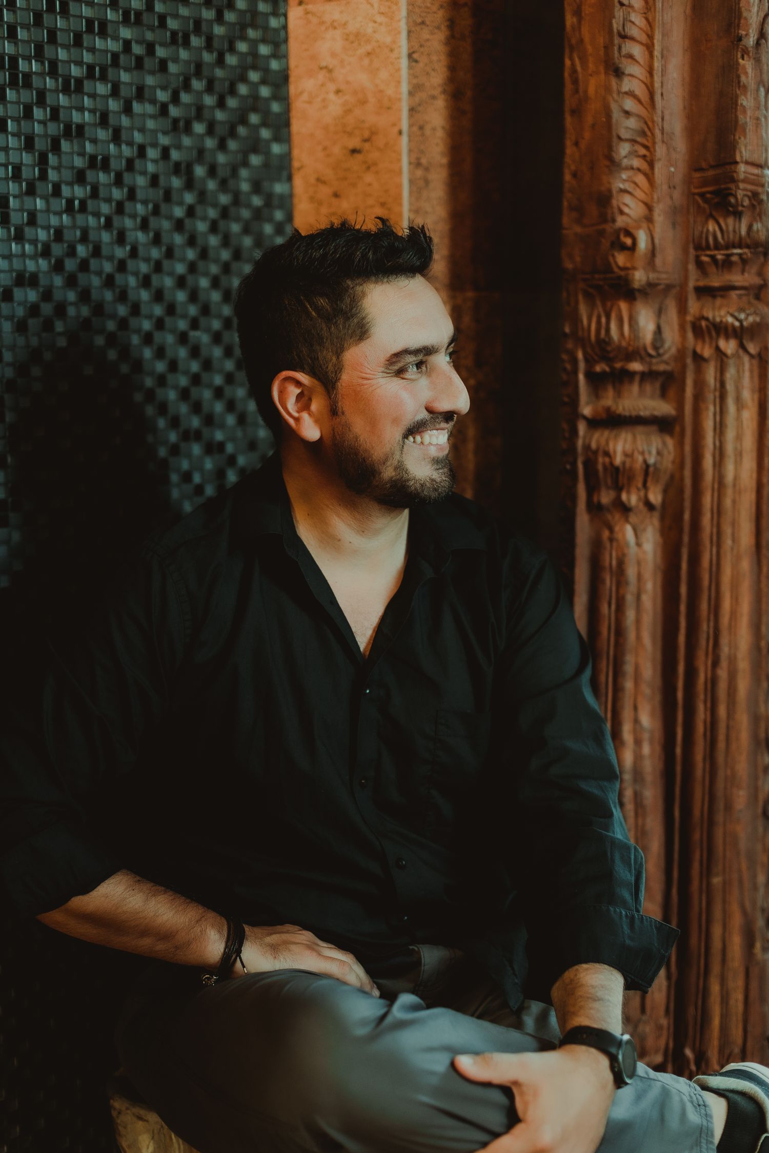 A person wearing black sits in contemplation near an ornate wooden doorway or wall panel in moody lighting.