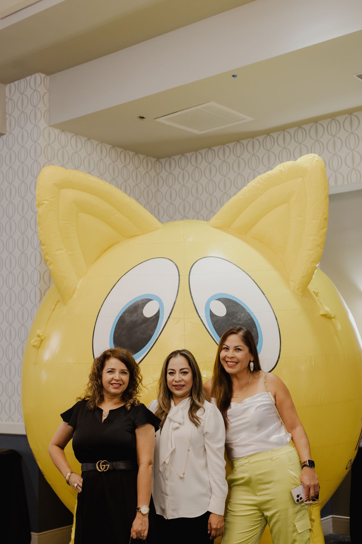 Three women pose together in front of a large yellow cartoon cat inflatable decoration with big blue eyes.