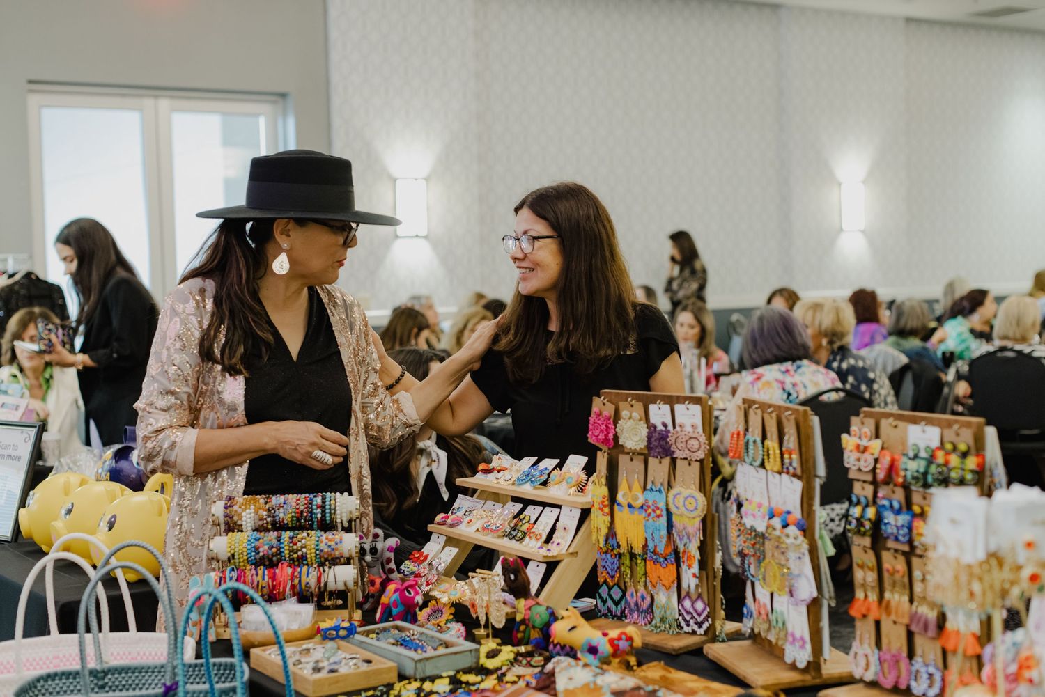 Vendors display colorful handmade jewelry and accessories at an indoor craft market while discussing their wares.