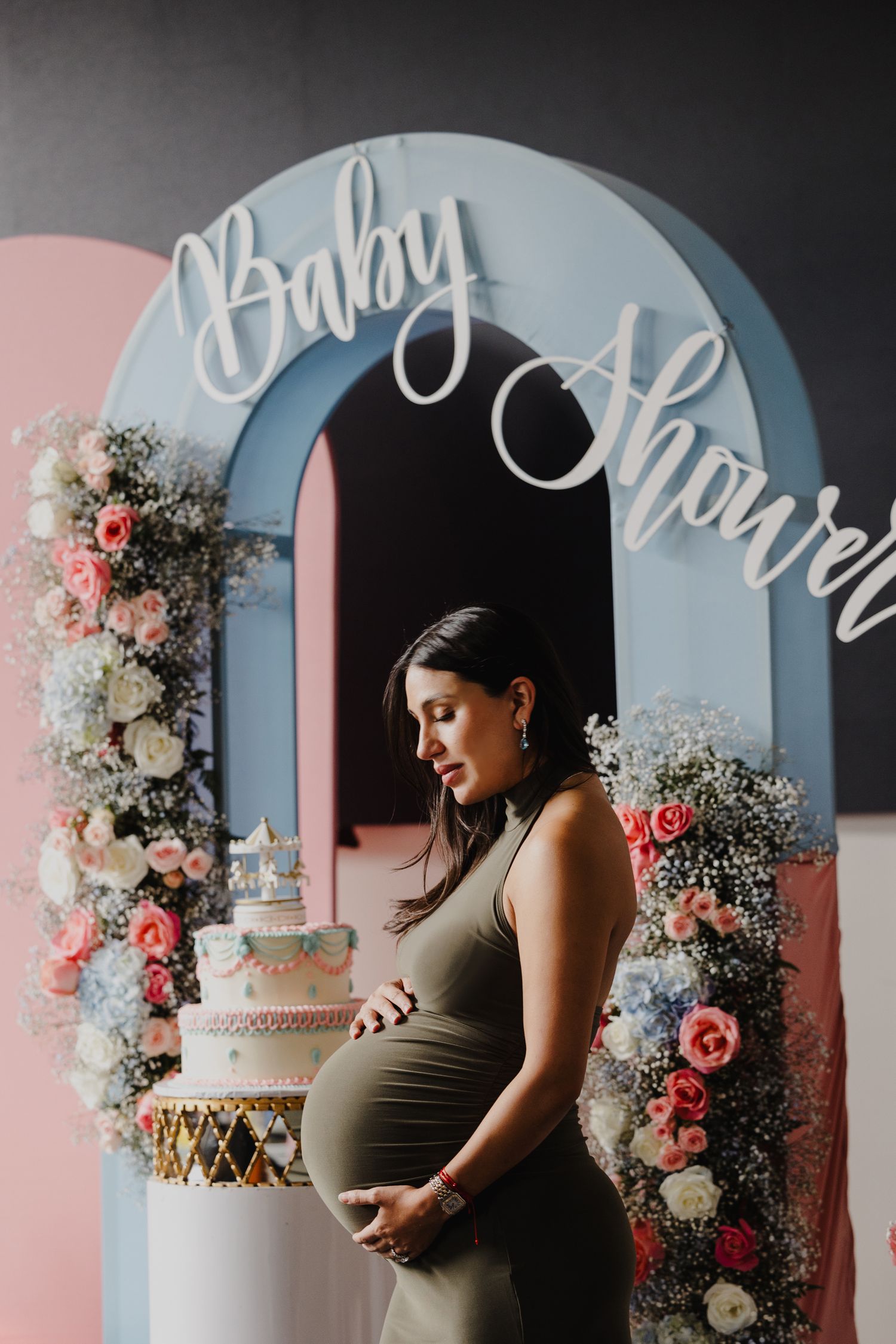 Beautiful baby shower setup with floral arch, elegant tiered cake, and expectant mother in brown dress against pink and blue backdrop.