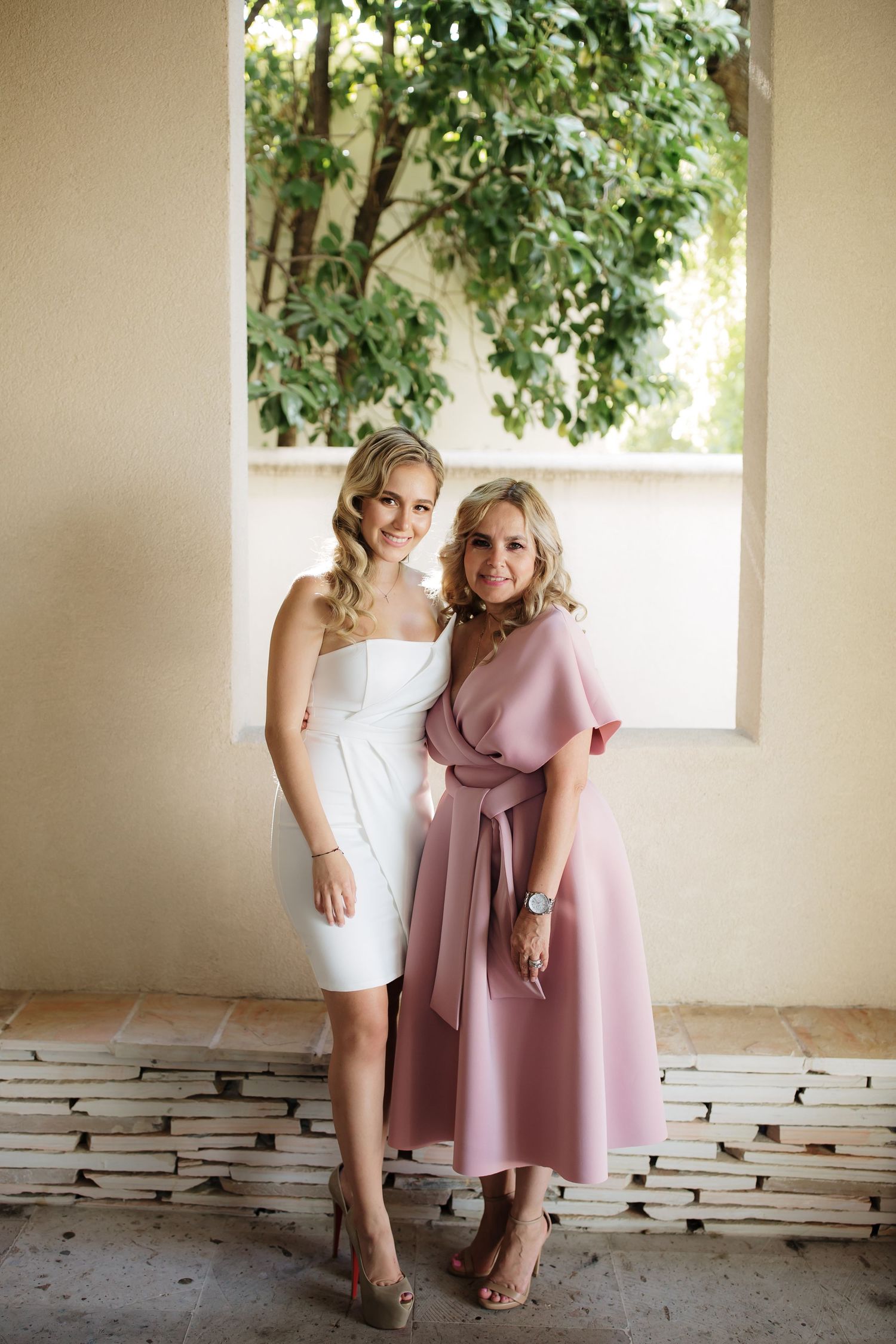 Two women pose elegantly in formal dresses, one in white and one in dusty rose, against a garden backdrop.