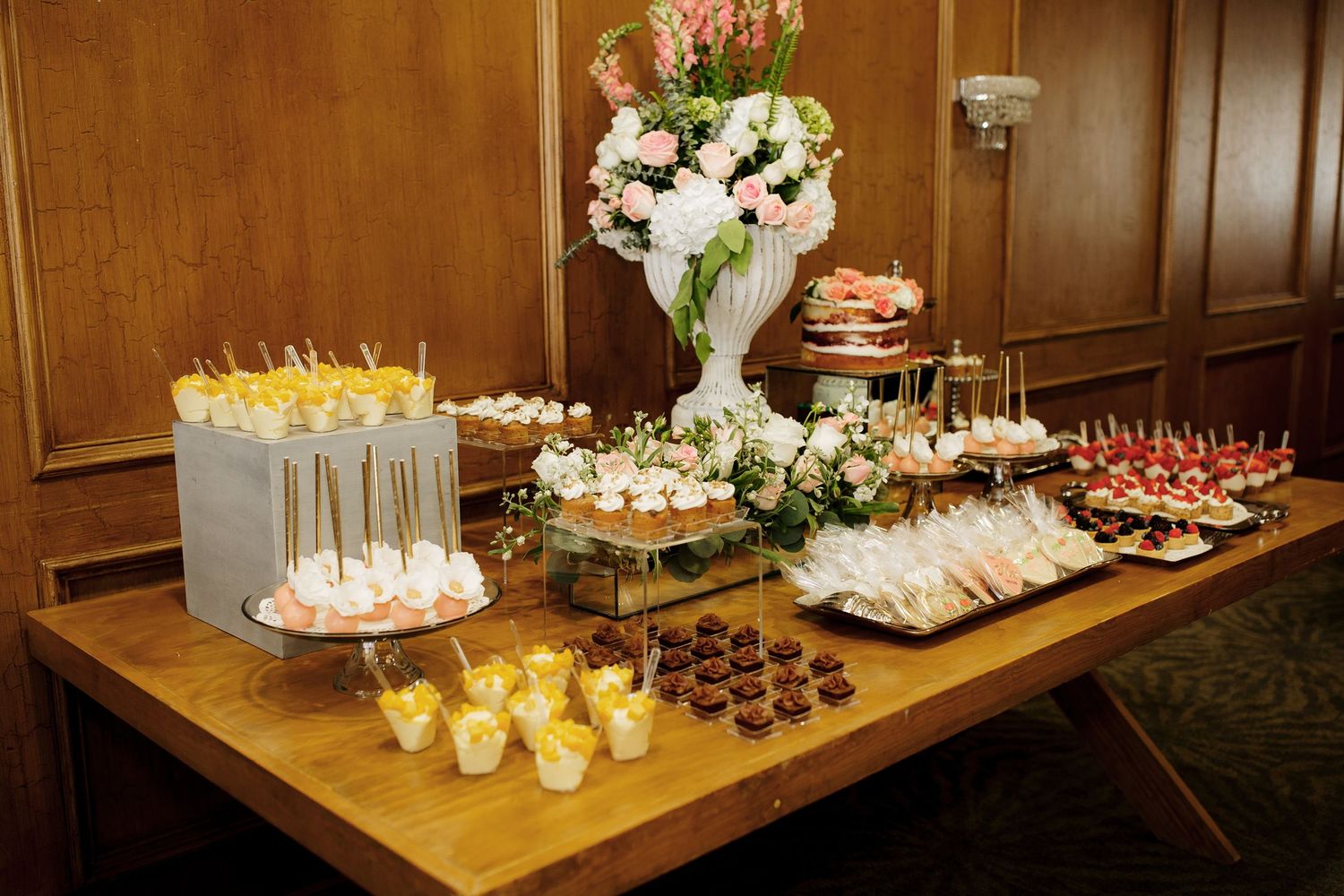 An elegant dessert table display with pastries, cakes, and a floral centerpiece arranged on a wooden buffet surface.