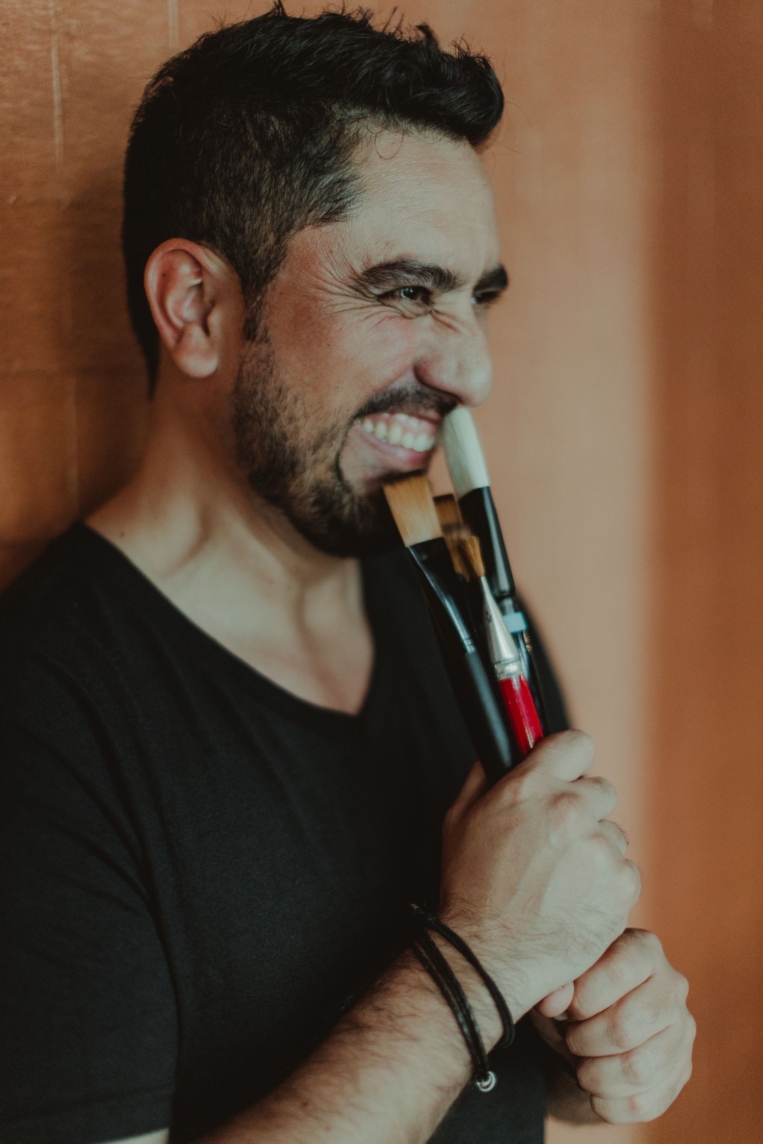 Close up portrait of someone smiling while holding makeup brushes against an amber-toned background.