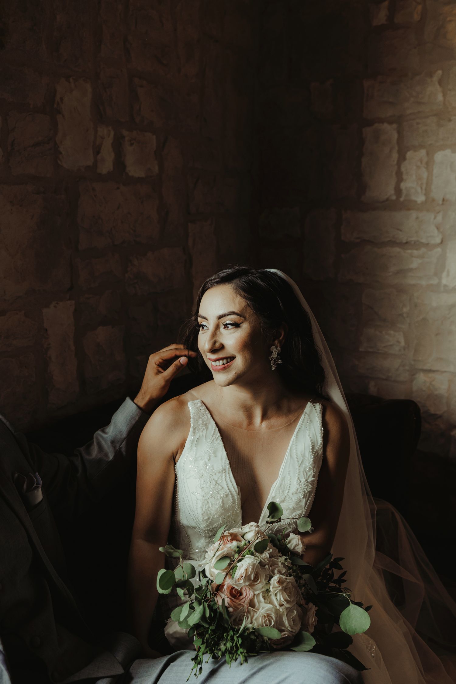 Moody portrait of bride in lace v-neck wedding gown holding delicate bouquet against stone wall backdrop.