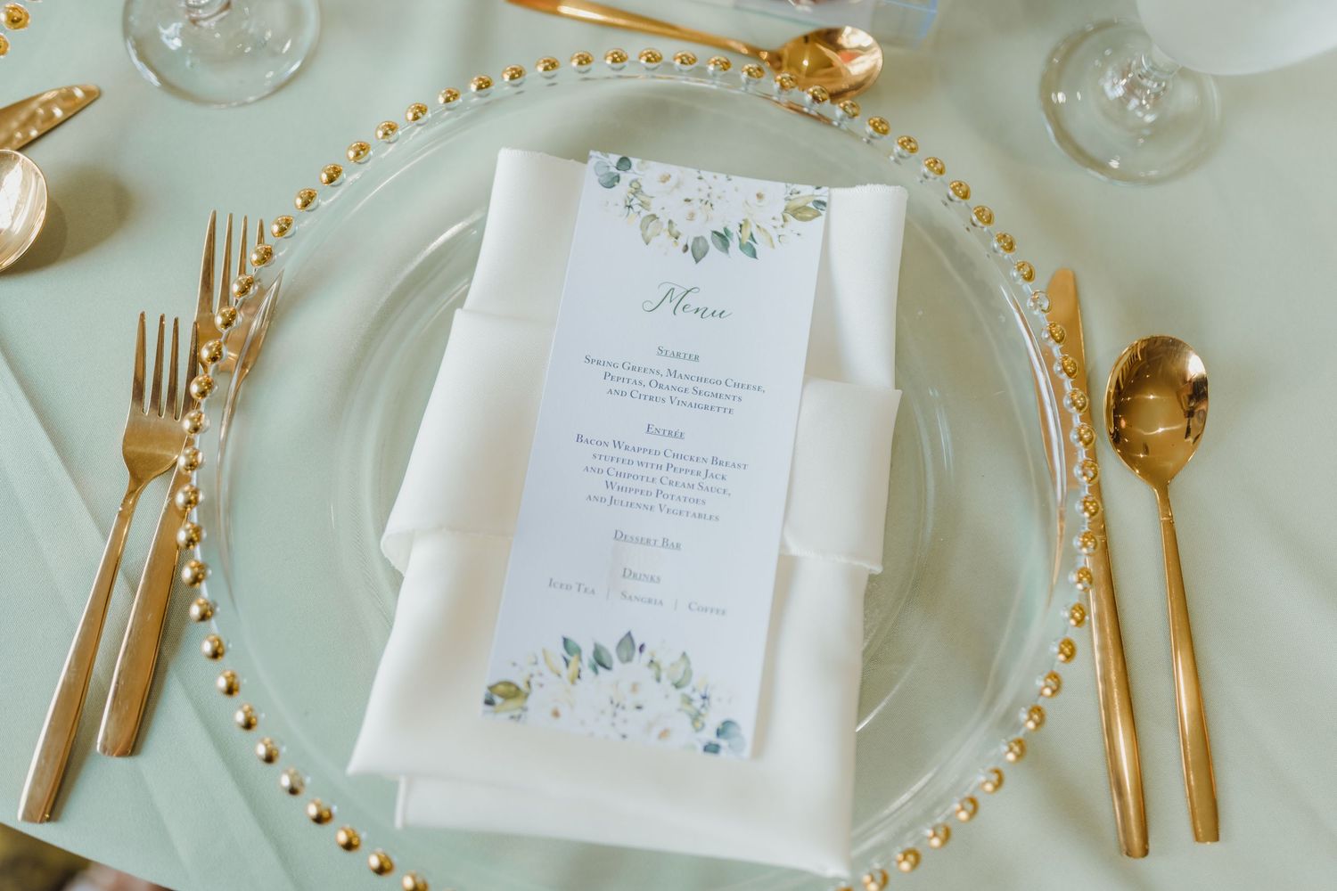 Elegant place setting with gold utensils, beaded charger plate, and floral wedding menu on white linen tablecloth.