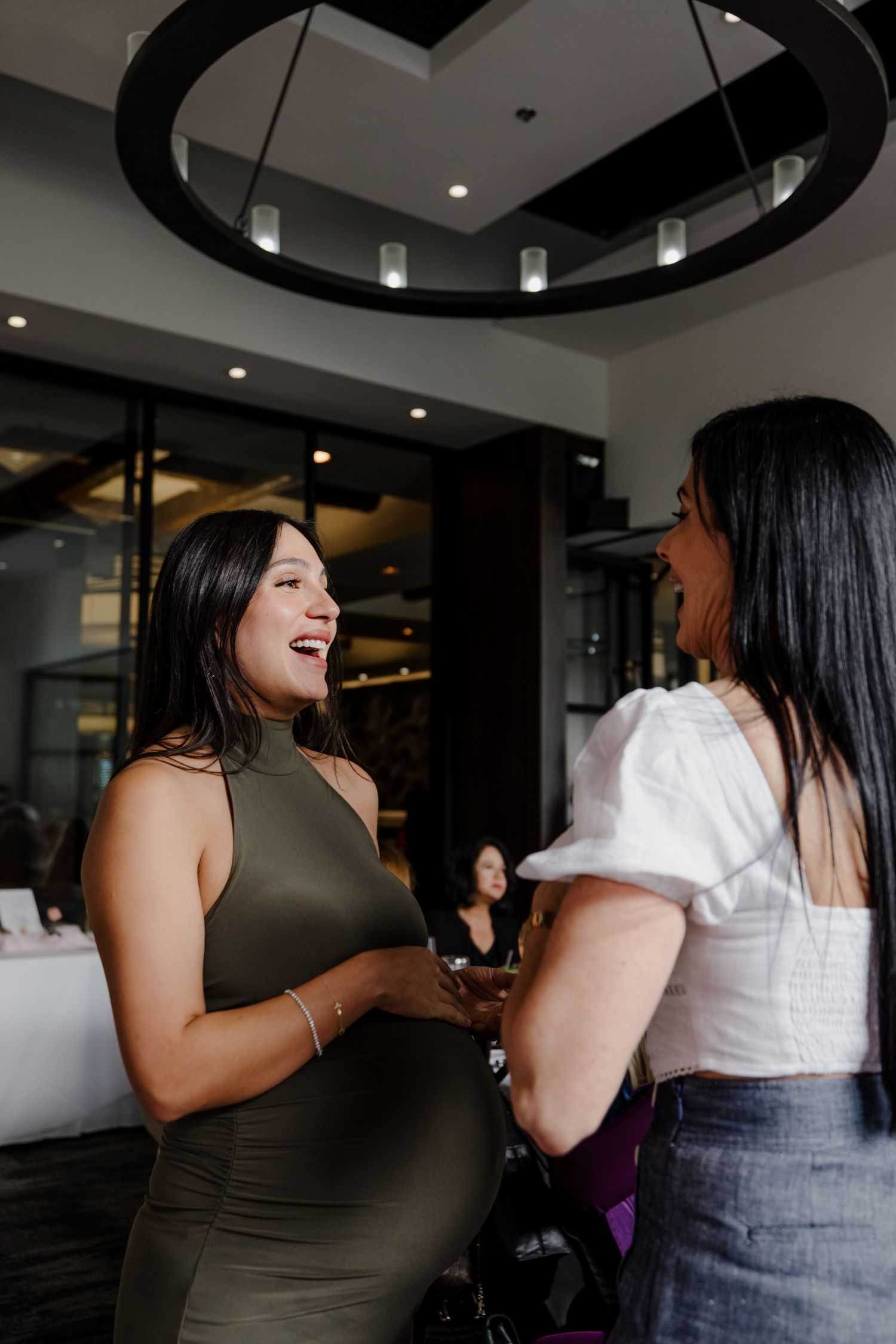 Two women in casual attire share a joyful conversation in an office space with dark circular lighting fixtures above.