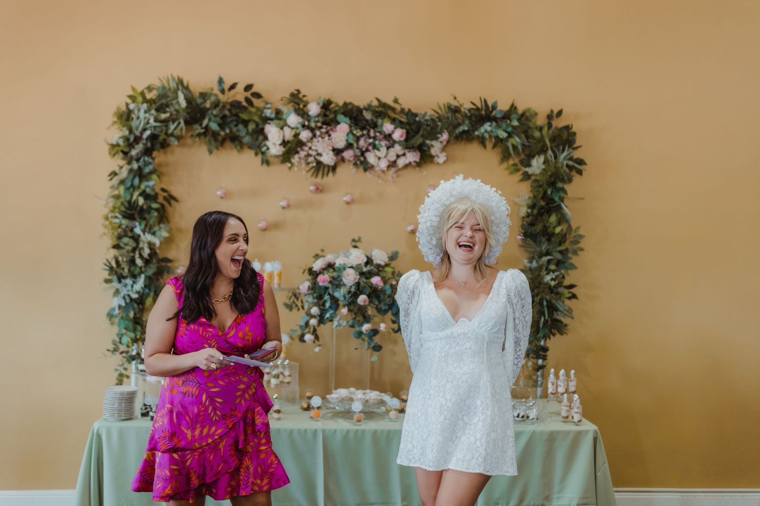 Two people stand by a decorated table with a green floral arch hanging above against a beige wall.