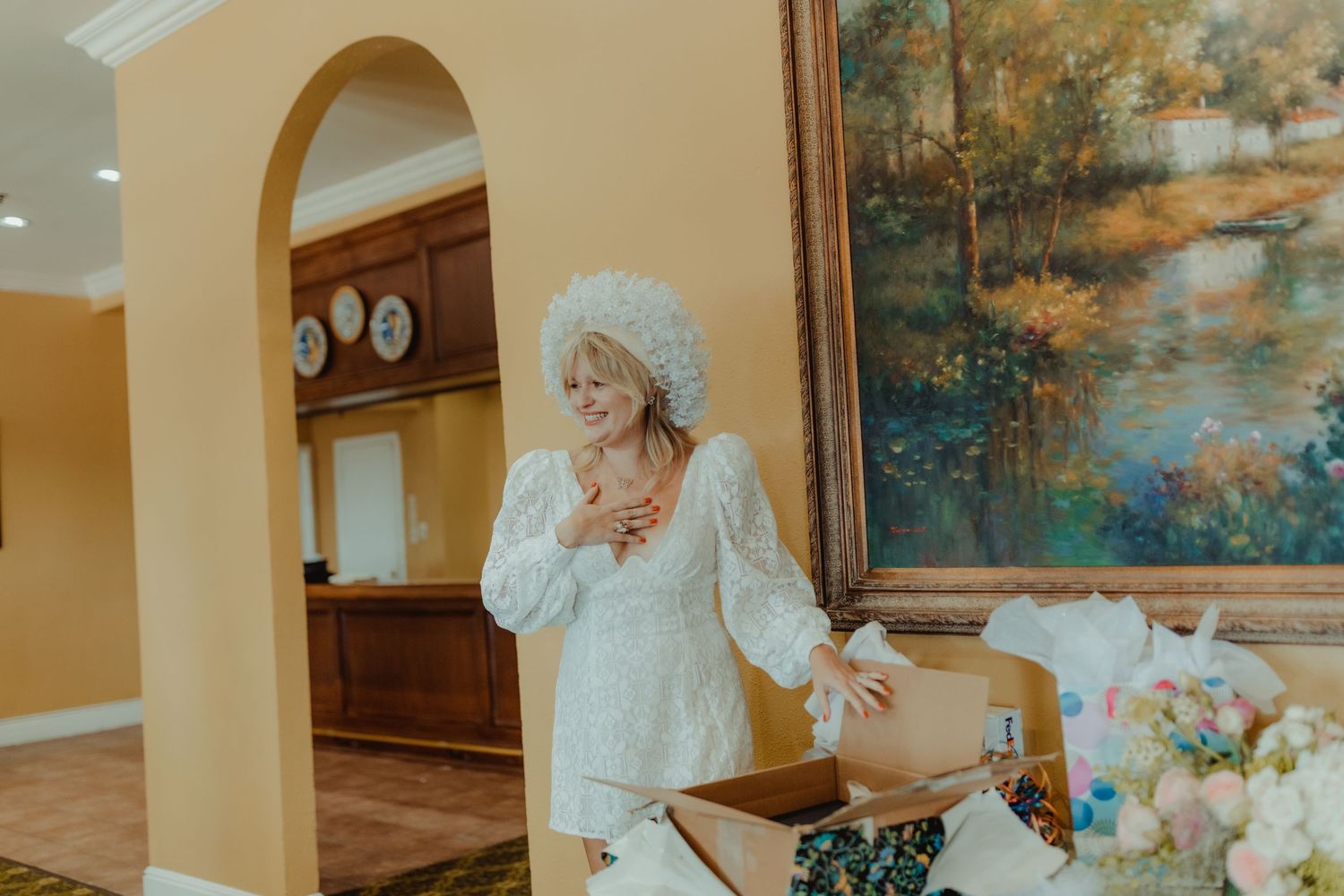 Person in period colonial style dress with white bonnet and lace top stands expressively in an elegant room with artwork.