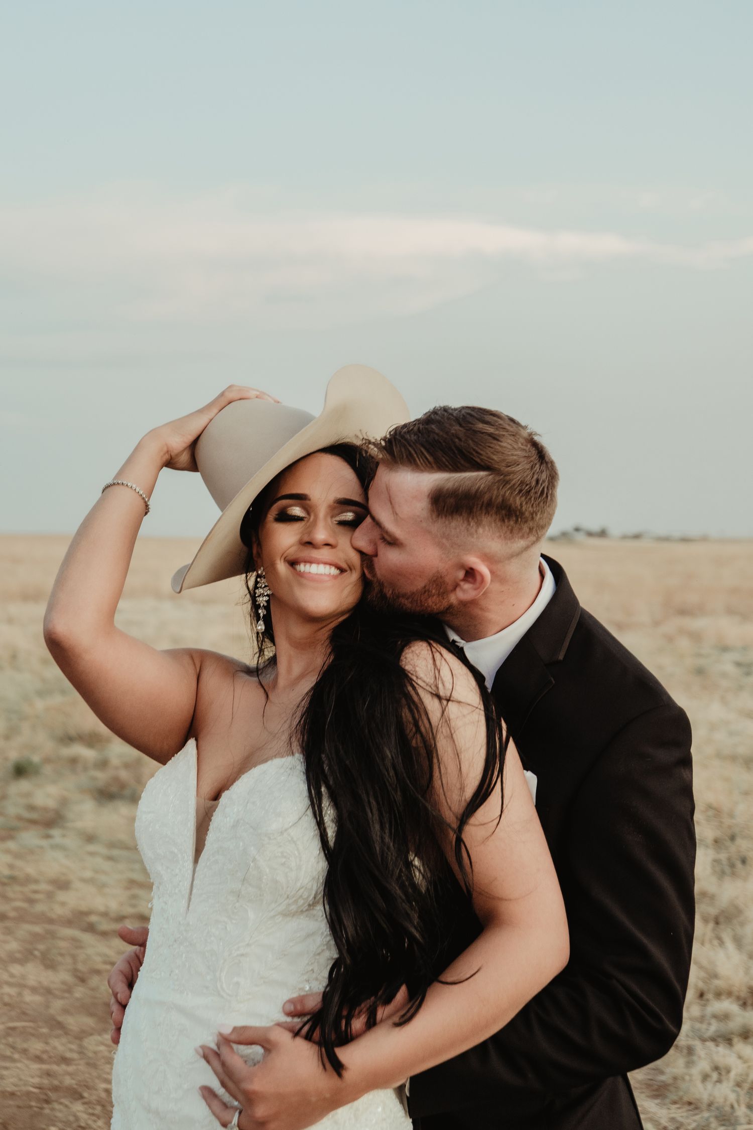 A happy couple embraces in an outdoor desert setting at sunset, with the bride wearing a cowboy hat and white dress.