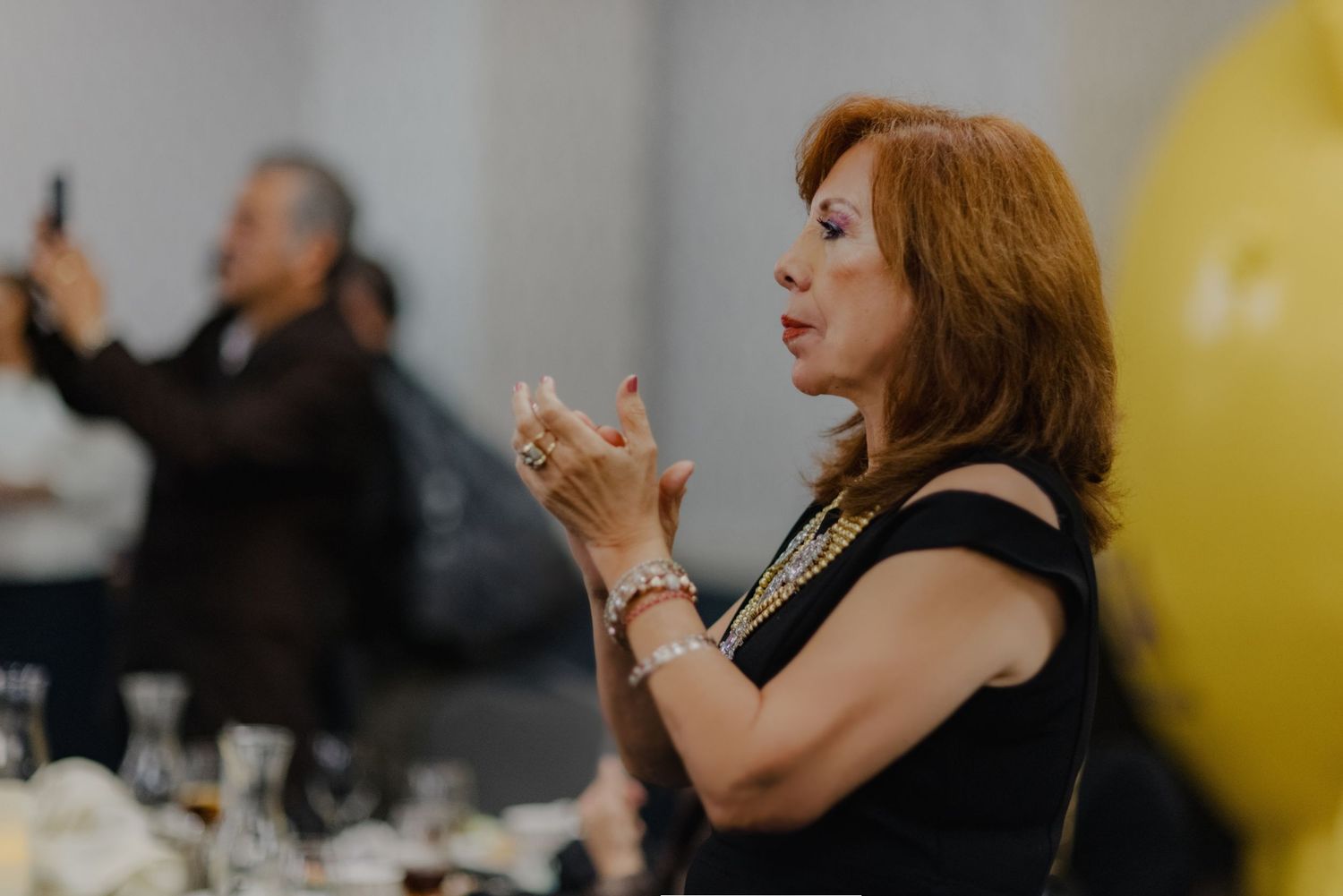 Woman in black sleeveless dress clapping at formal event with balloons in background.