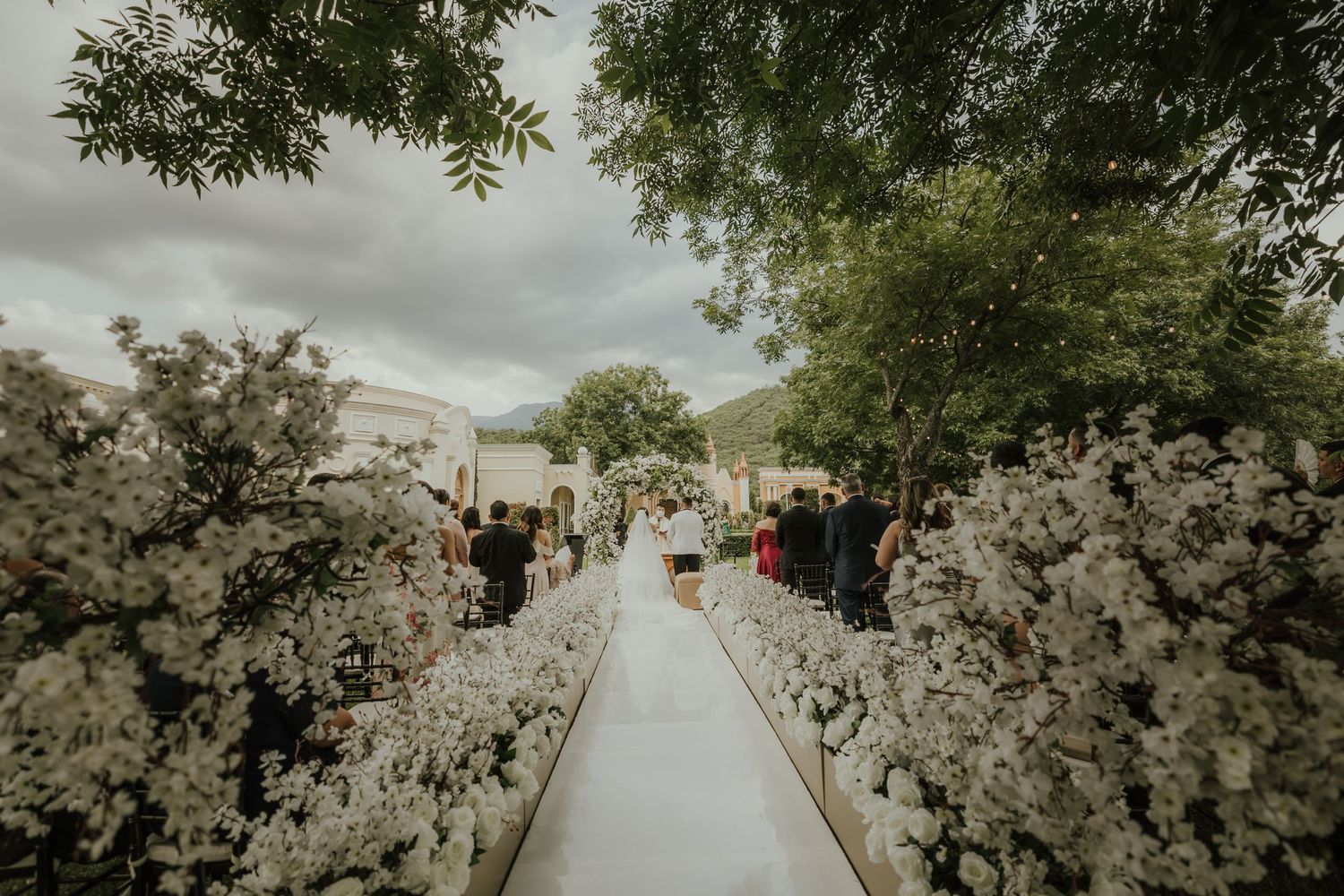 White floral arrangements line both sides of an outdoor wedding aisle with a dramatic cloudy sky backdrop.