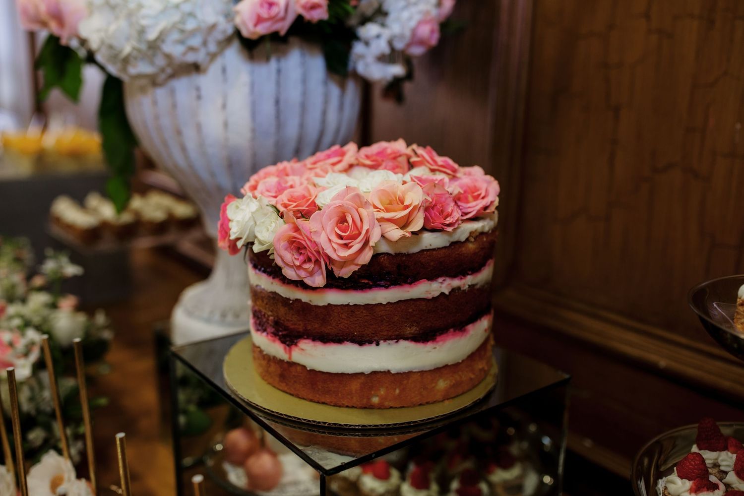 A rustic naked wedding cake topped with fresh pink roses and white cream filling sits on a glass pedestal.
