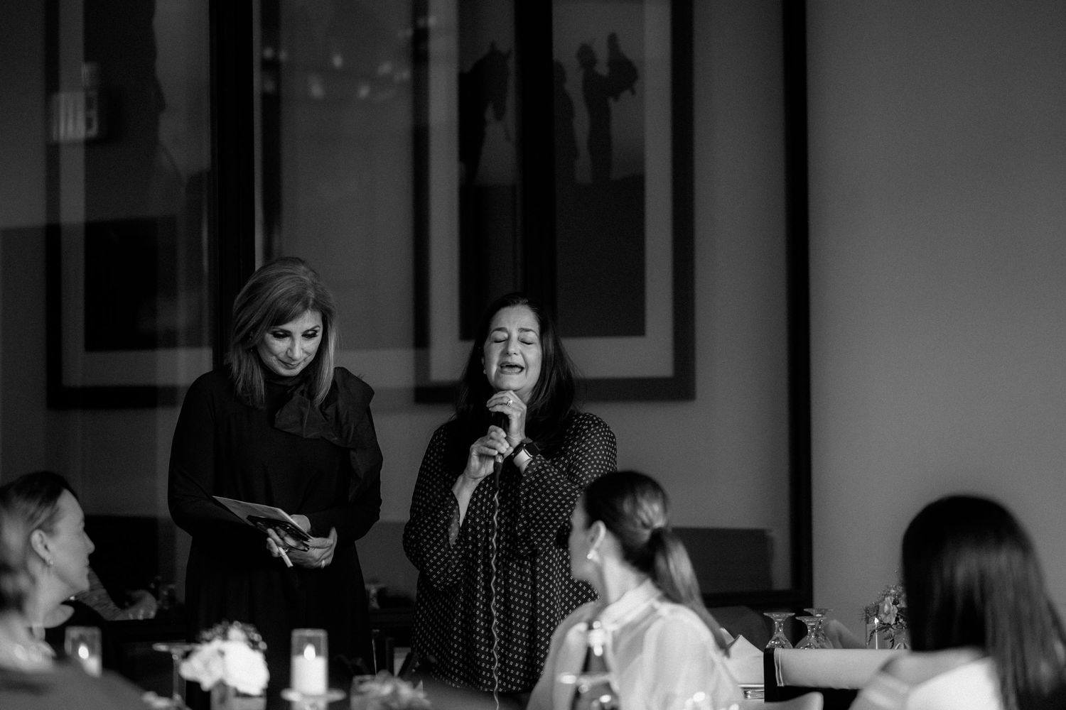 A black and white candid moment captured during a gathering at an indoor venue with people seated at tables.