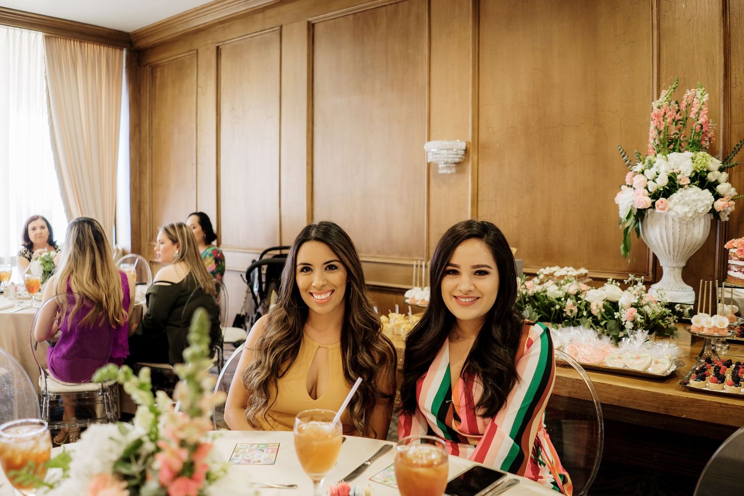 Two women in colorful dresses sitting at an elegant dining event with floral arrangements and guests in the background.
