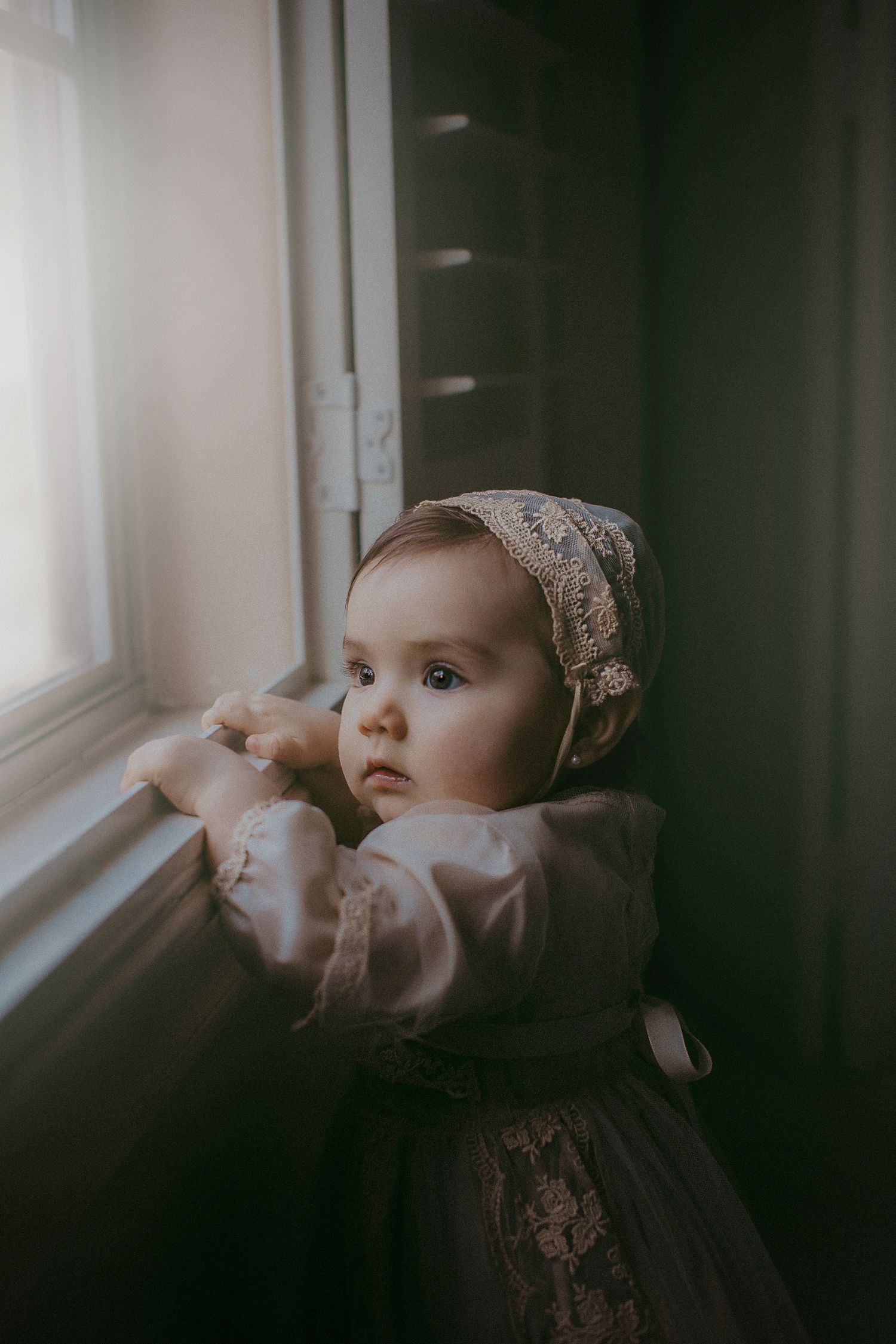 A young child in vintage dress and bonnet gazes thoughtfully out of a sunlit window.