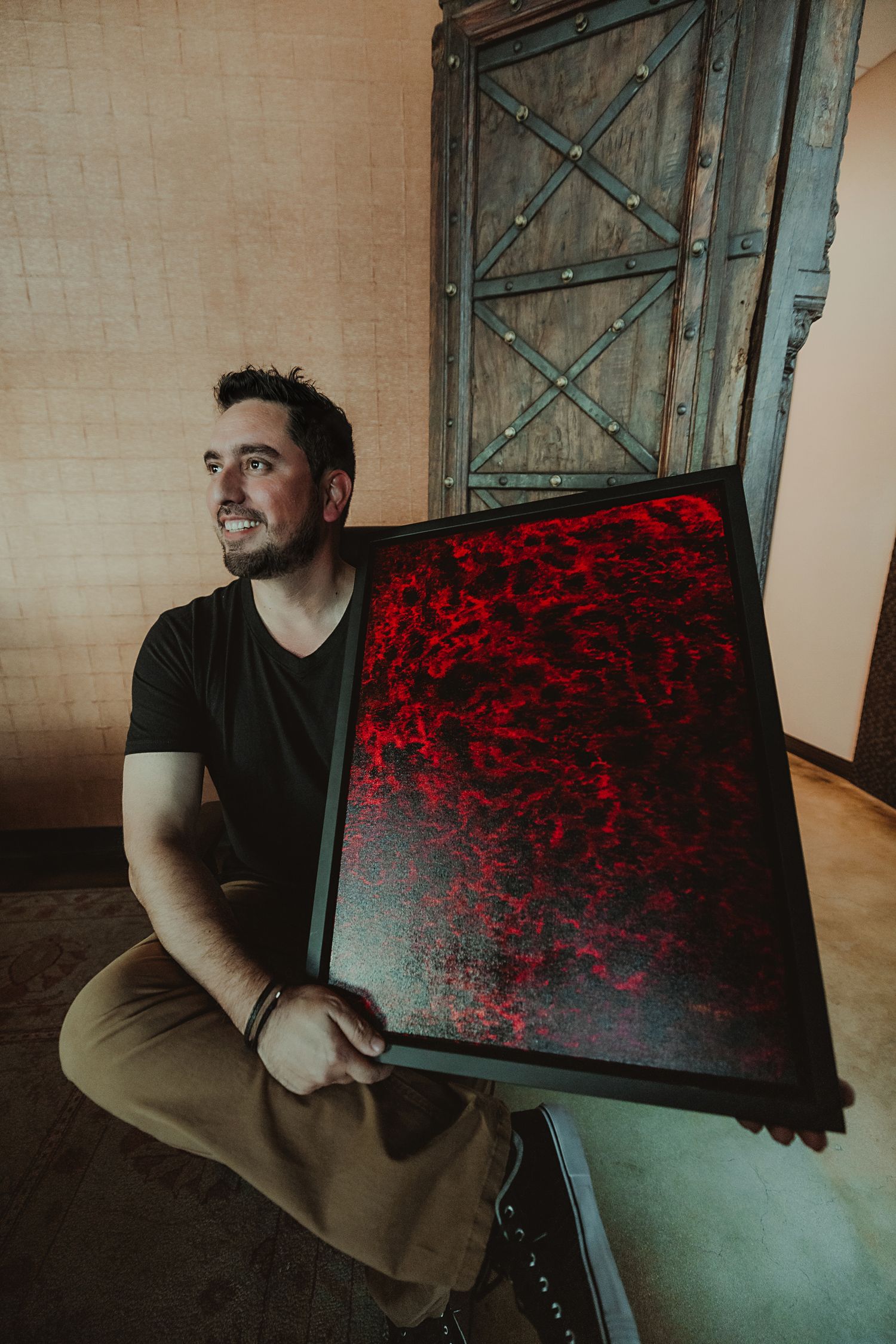 Artist poses with abstract red and black painting displaying on textured metal-framed canvas.
