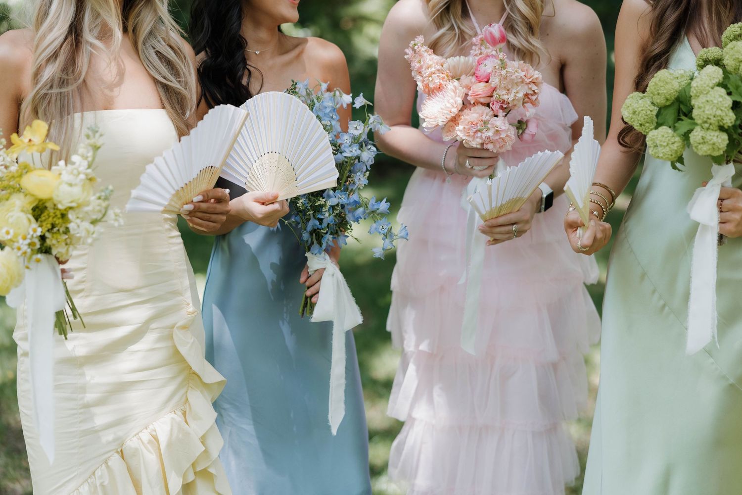 Bridesmaids in pastel dresses holding delicate bouquets and fans at an outdoor wedding.