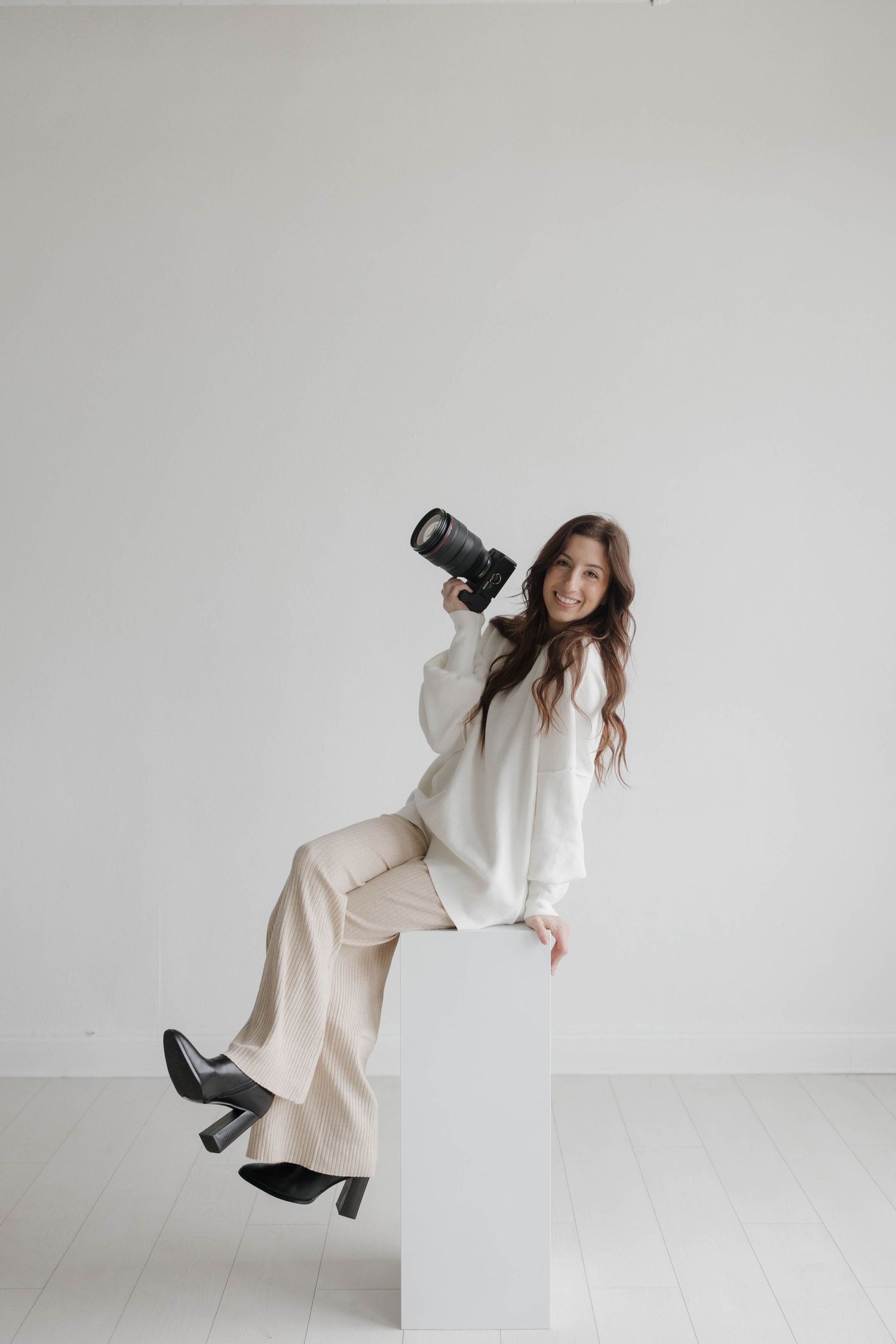 Professional photographer in white outfit sitting on cube holding camera in studio setting.