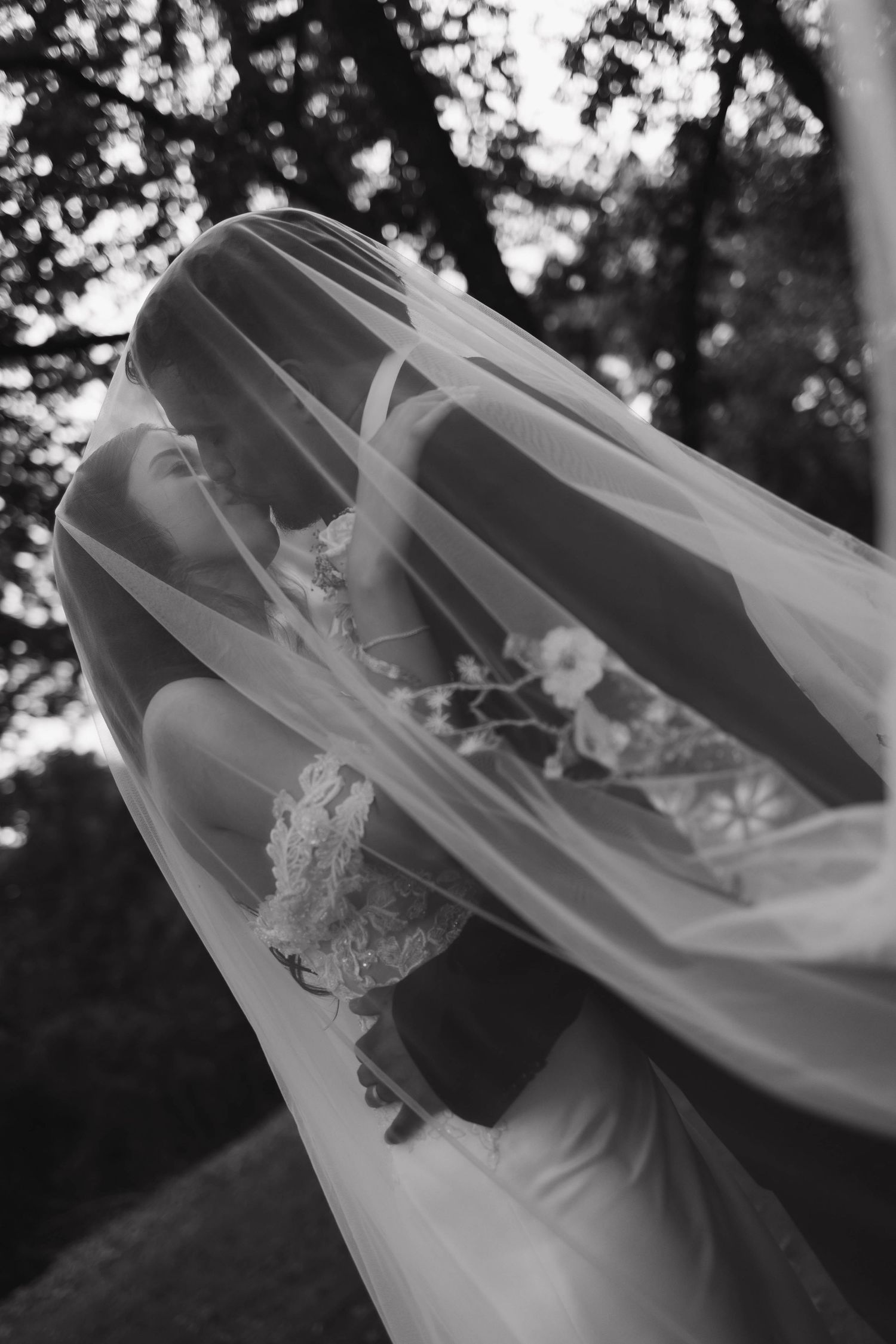 Artistic black and white photo of bride with flowing veil against trees.