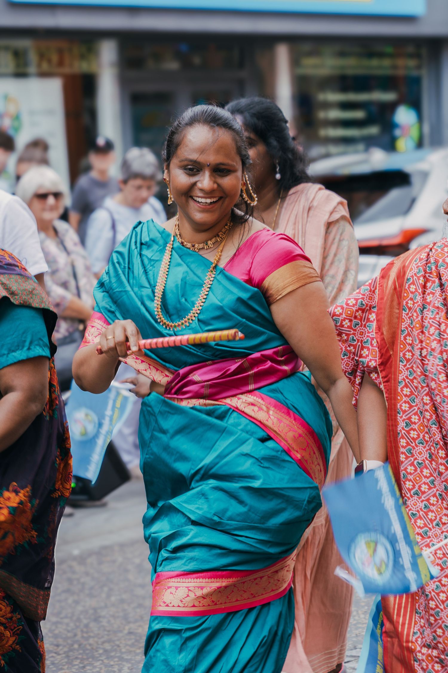 A smiling person in a vibrant turquoise and pink silk sari with gold jewelry dancing at a cultural celebration.