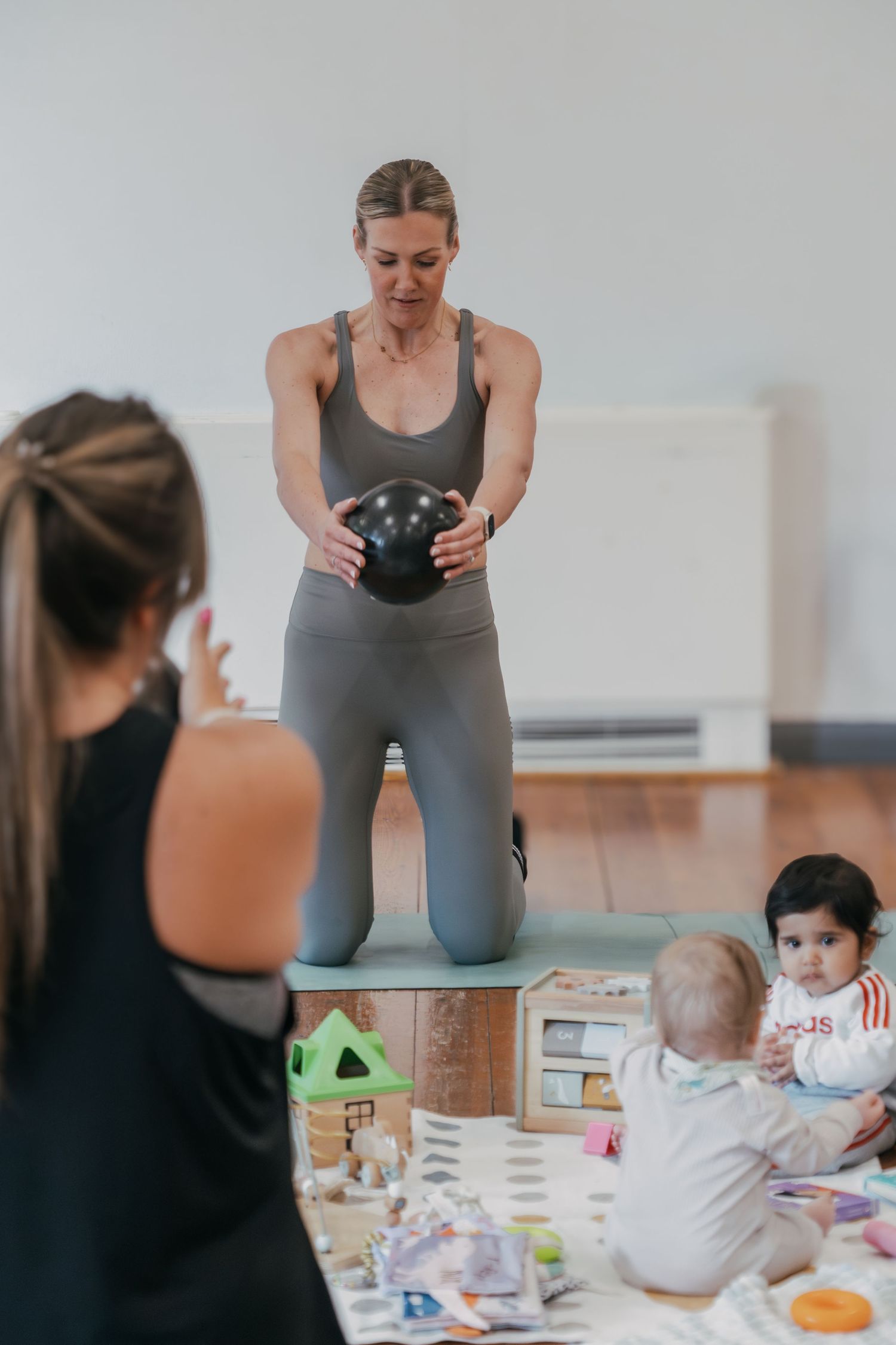 A person exercising with a medicine ball while toddlers play with toys on the floor in a home workout space.