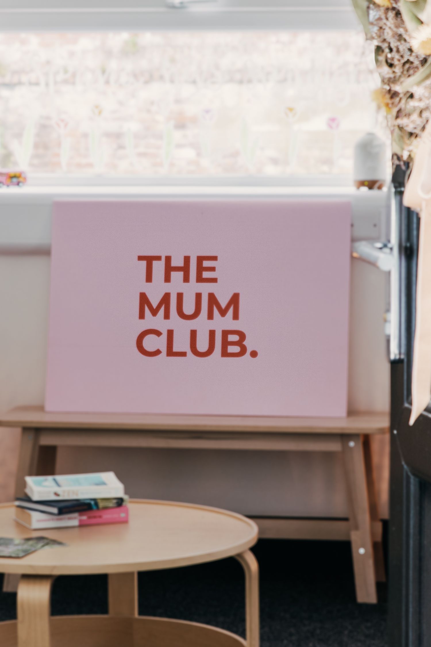 A pink sign with red text reading 'THE MUM CLUB' displayed on a wooden table alongside some stacked books.