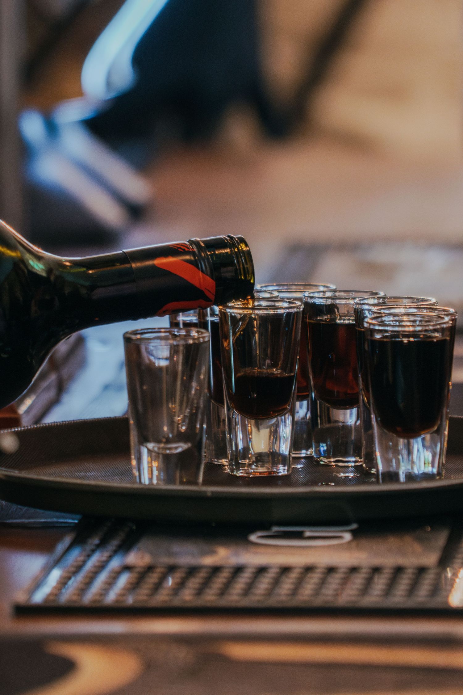 Dark liquor being poured into shot glasses on a serving tray in a dimly lit setting.