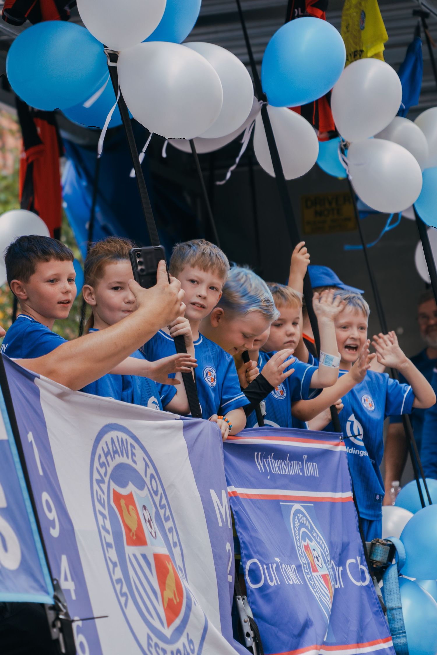 Young sports fans cheer excitedly behind team banners with blue and white balloons at a sporting event.