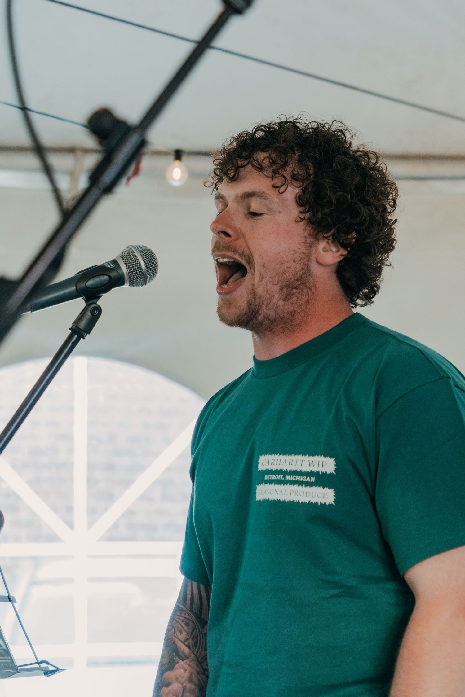 A performer in a green t-shirt sings passionately into a microphone during an outdoor performance under a tent.