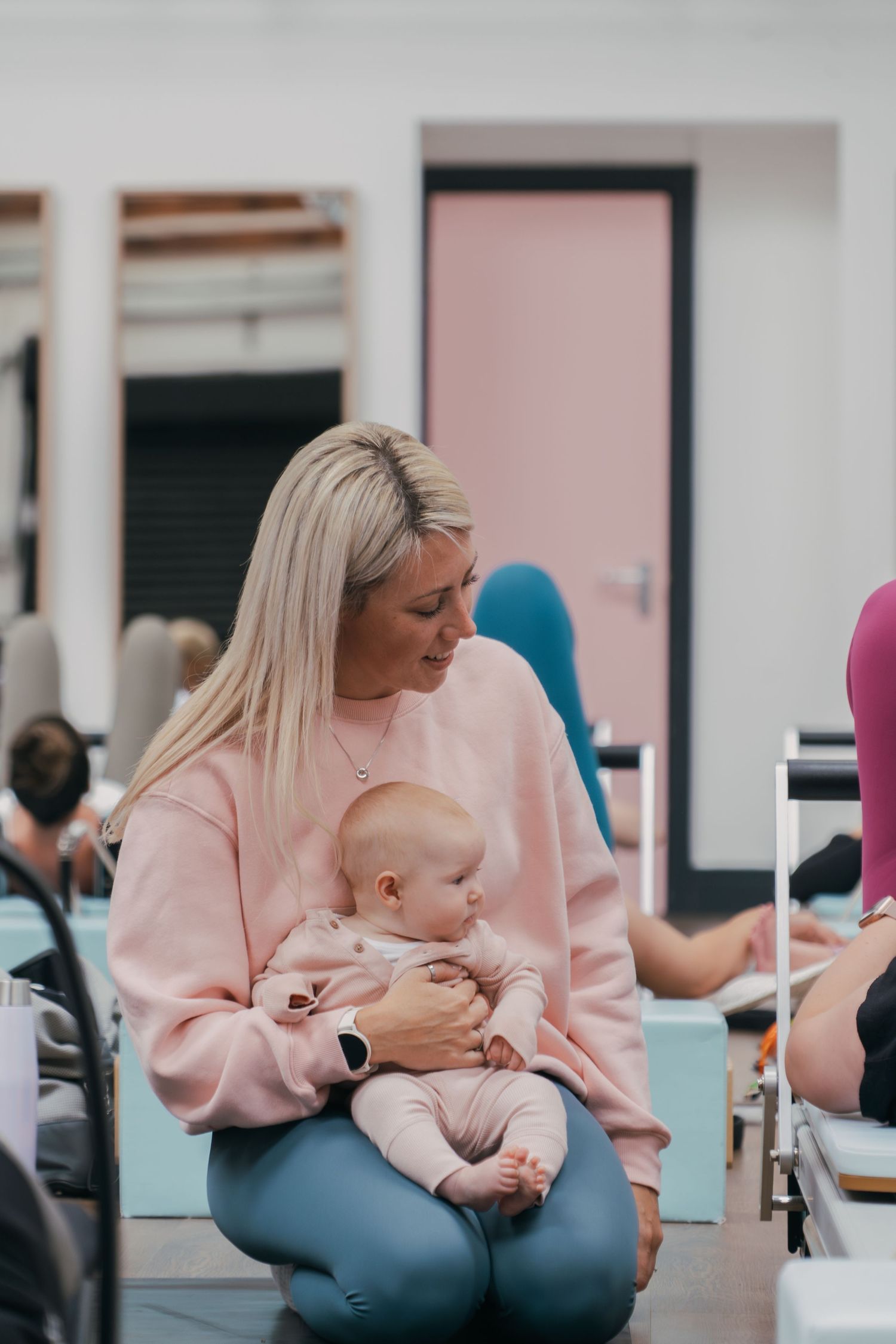 Parent wearing pink sweater and blue jeans holds baby in seated position during indoor gathering.