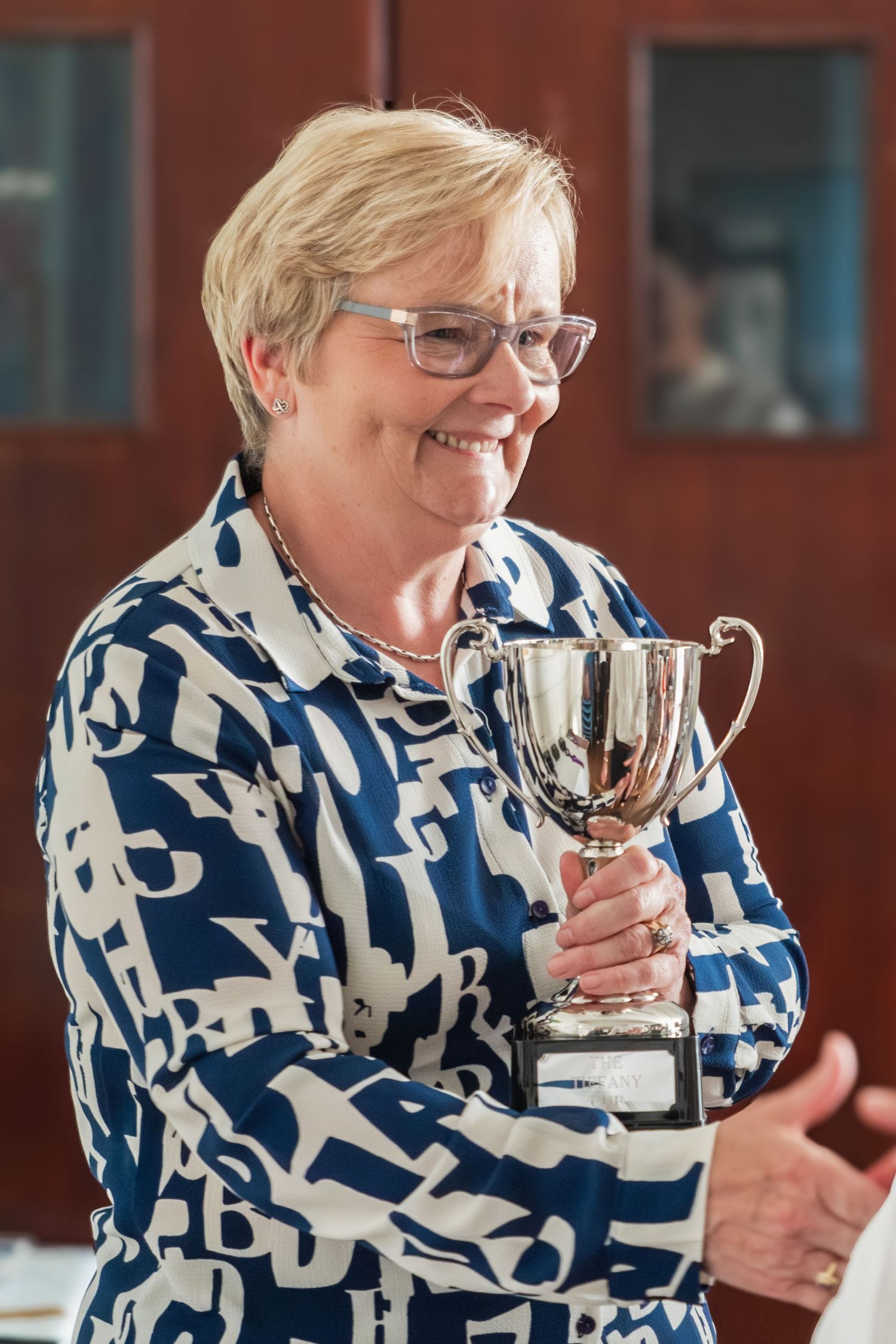 A winner displays a silver trophy cup while wearing glasses and a patterned blue and white shirt.