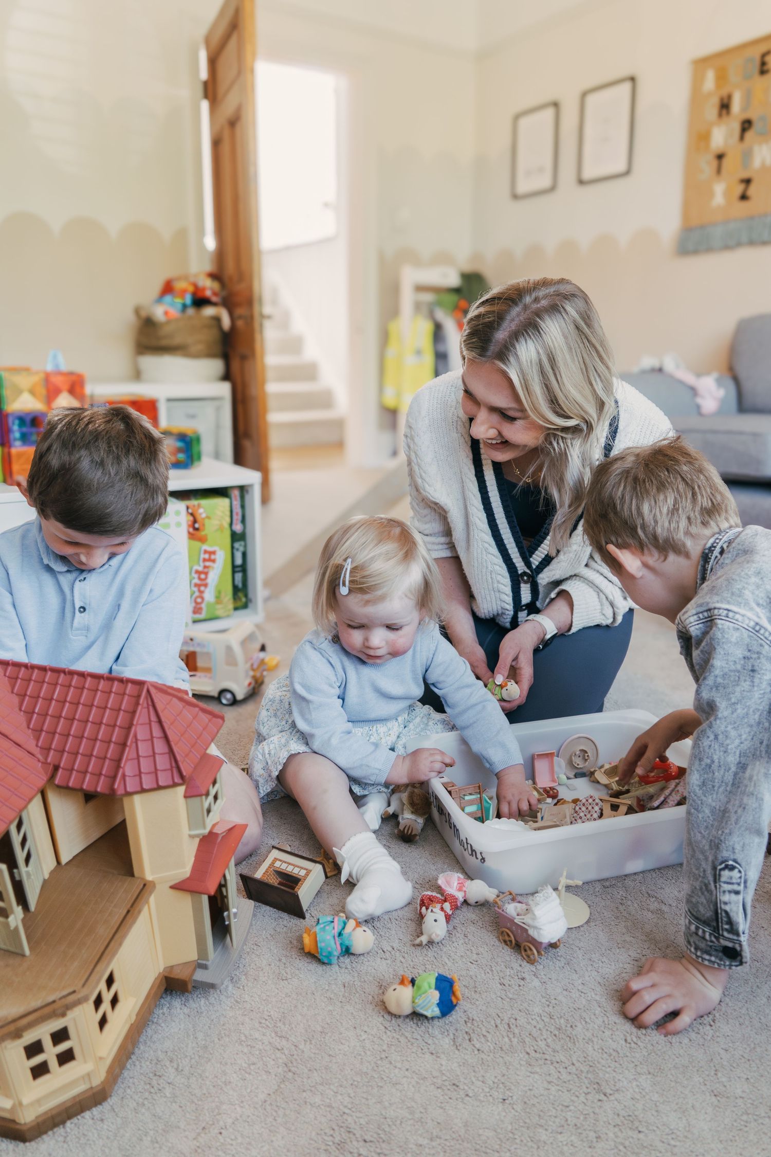 Kids playing with wooden dollhouse while their caregiver joins in on floor of bright living room.