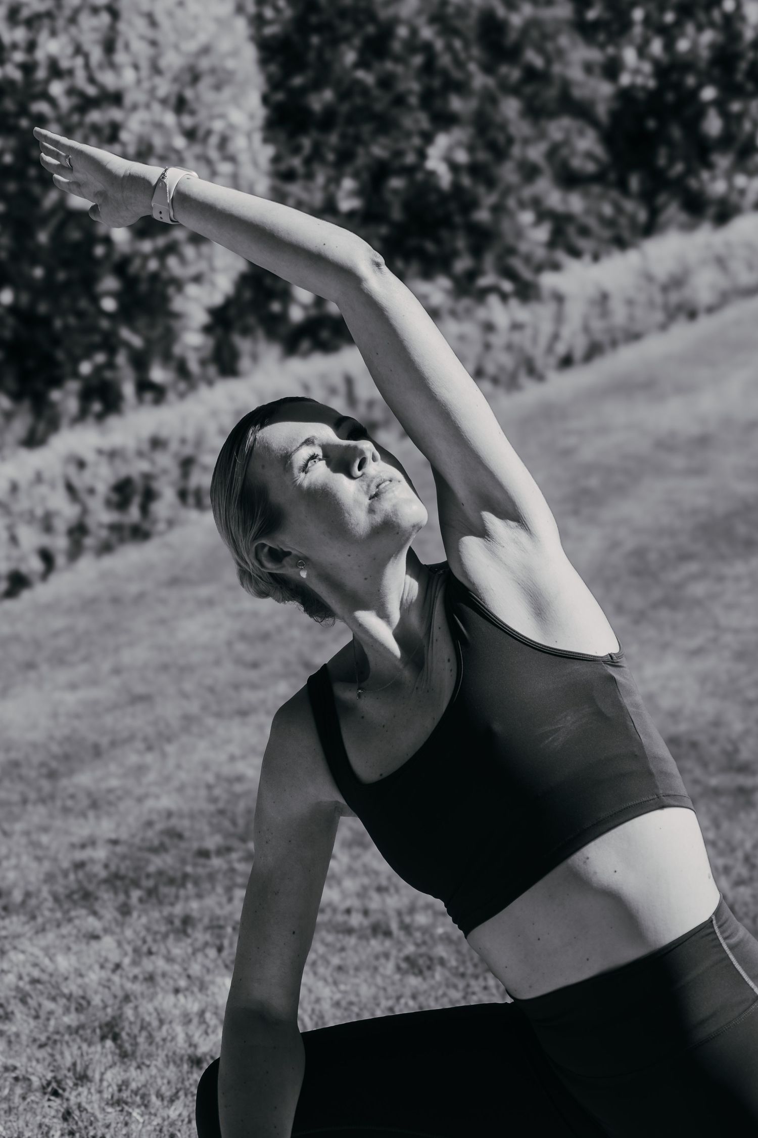 Person in black athletic wear performs a side stretch yoga pose on grass in a black and white photograph.