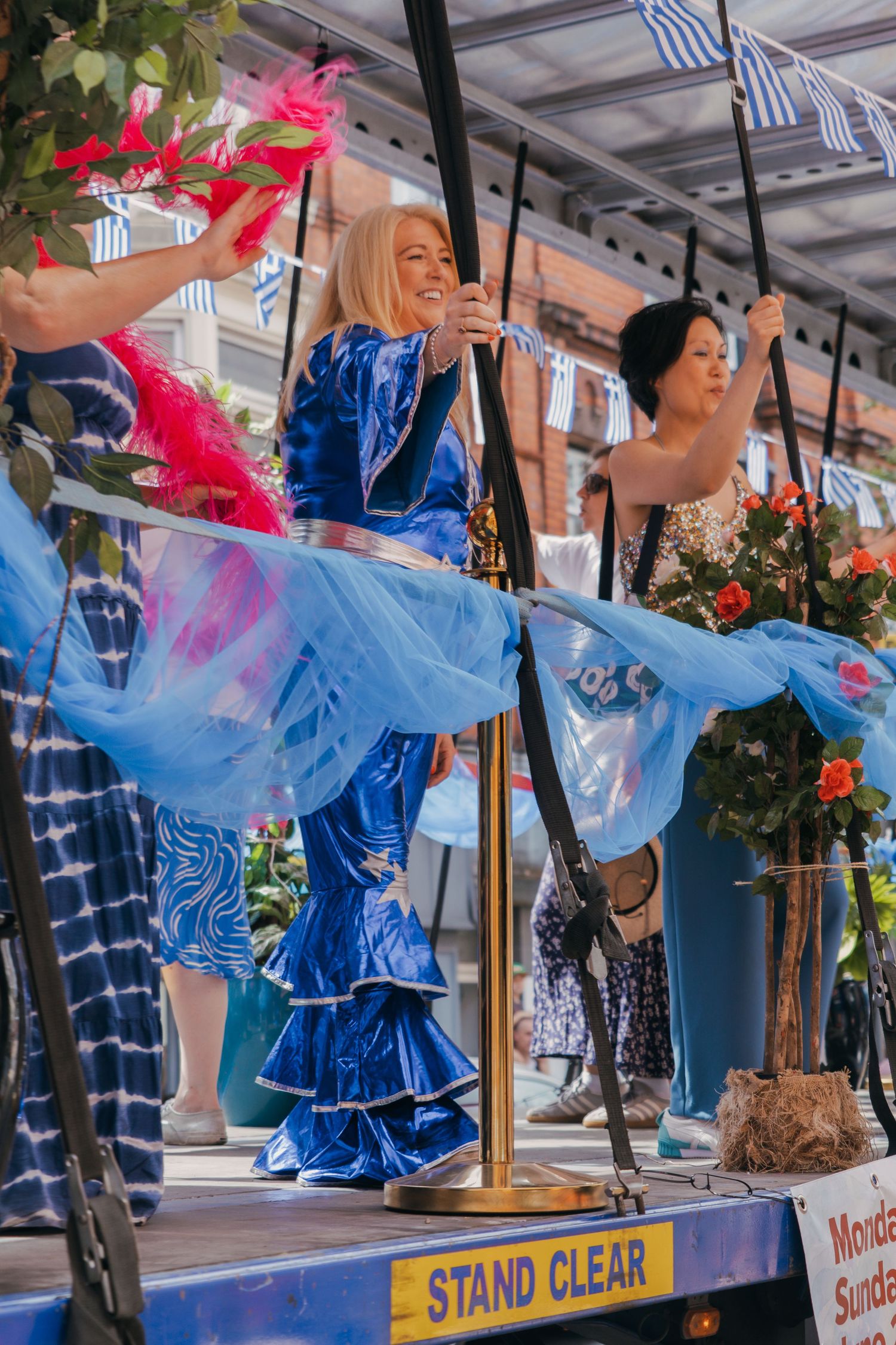 Performers in vibrant blue costumes dance and sing on a festival float decorated with colorful fabrics and flowers.
